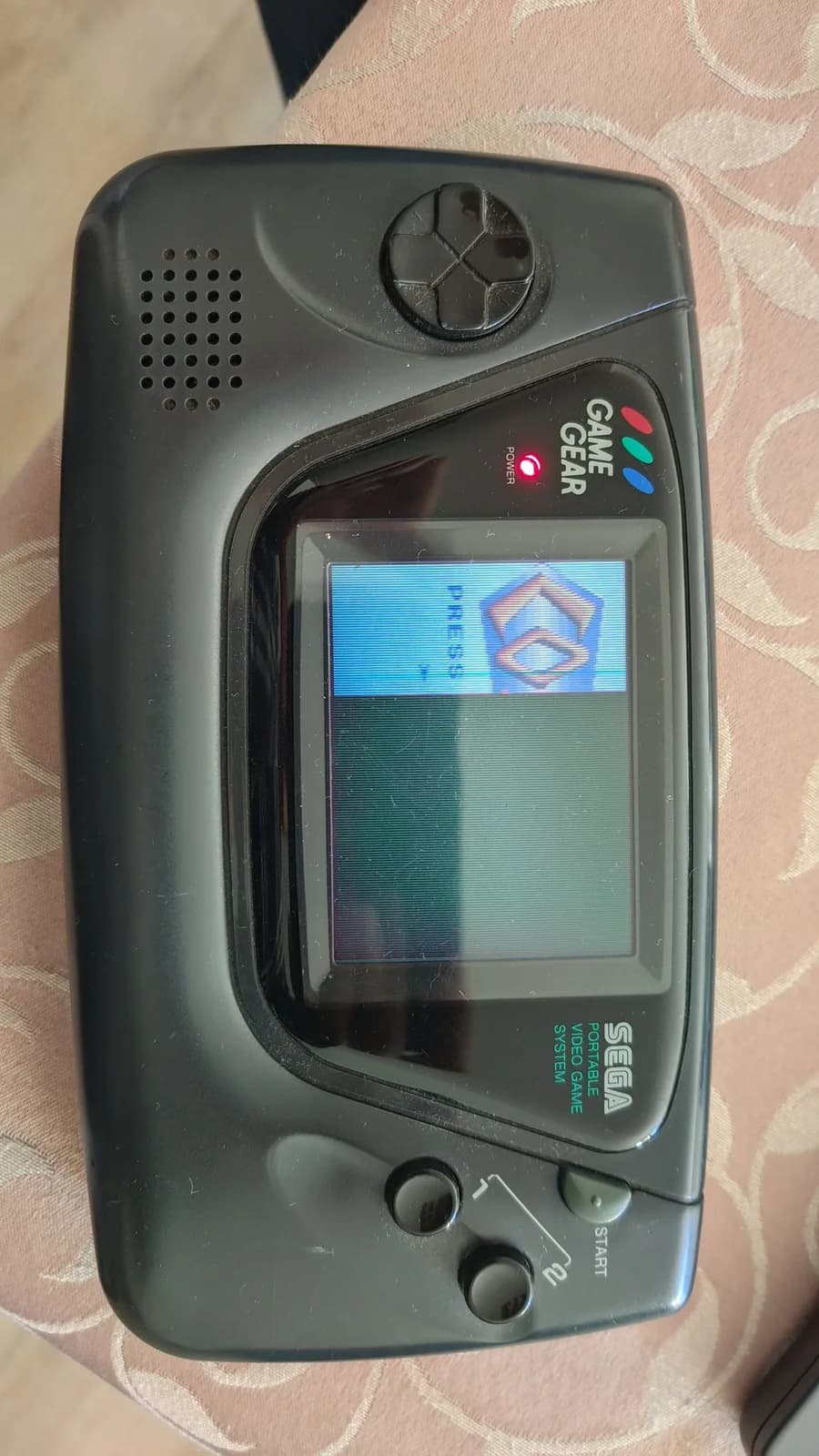Black Sega Game Gear powered on with its original, failing stock LCD: the Columns title screen is only partly visible because roughly two-thirds of the screen is dead and not displaying, the fault that prompted the IPS screen mod. Speaker grille top left, START button bottom right, resting on a patterned cloth.