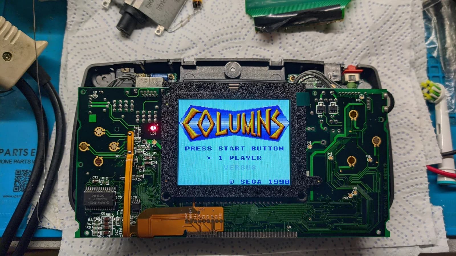 Sega Game Gear mainboard seen from the front after the IPS install (before the later accident), the working IPS screen showing the Columns title screen (PRESS START BUTTON, 1 PLAYER, Sega 1990), with the orange IPS flex cable routed across the green PCB.