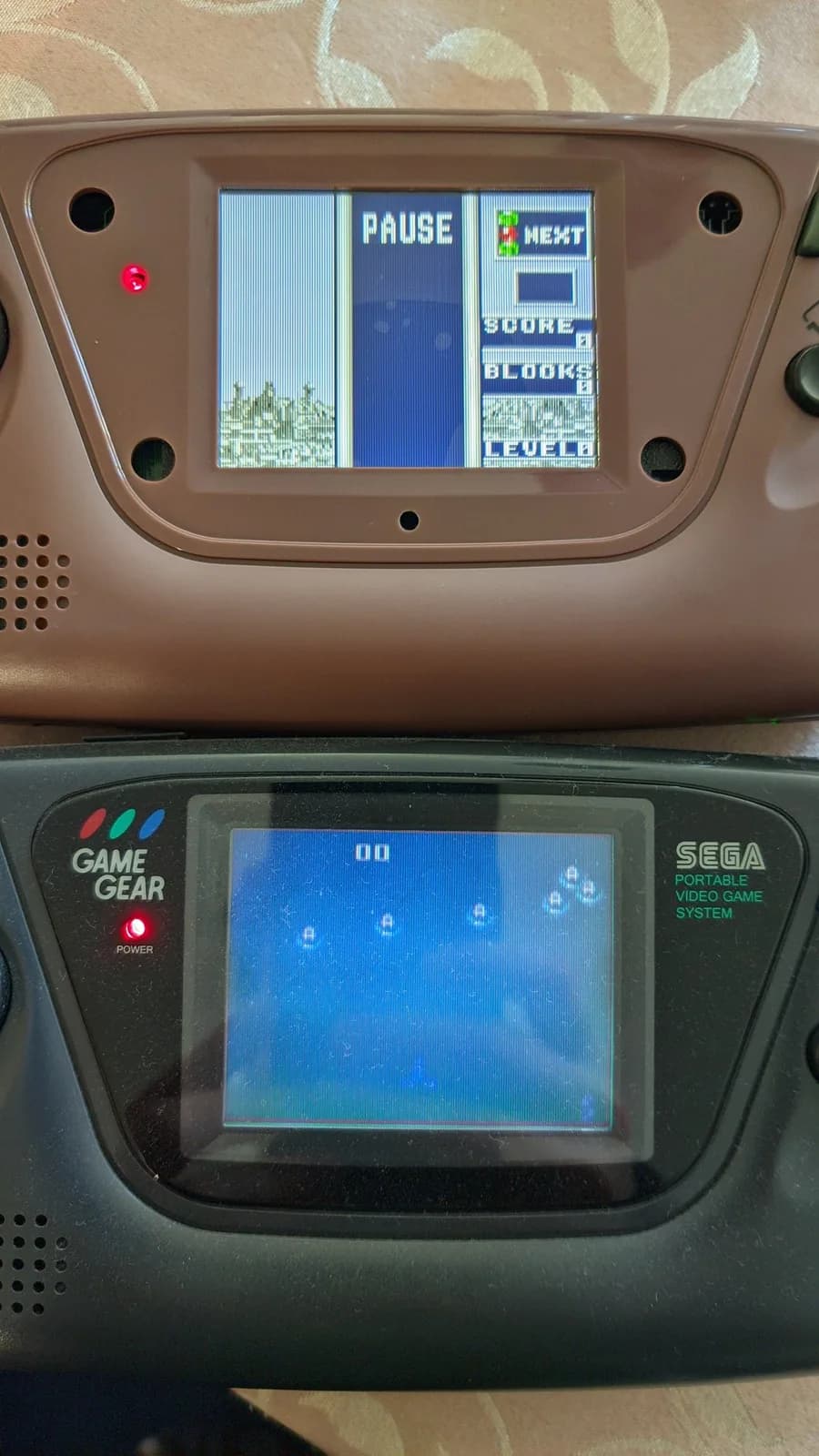Two Sega Game Gear handhelds stacked vertically; top unit shows IPS screen displaying pause menu with score/block/level; bottom unit shows stock screen with blue display.