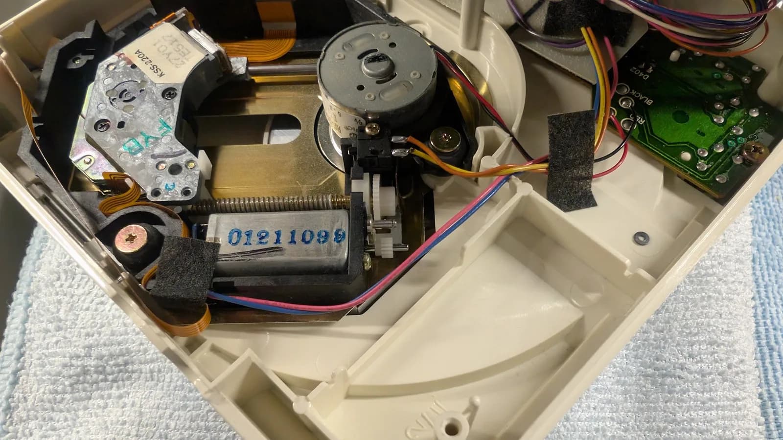 PC Engine CD-ROM2 internal mechanism with the laser assembly, motor, PCB and wiring on the white plastic chassis, showing the shaft where the old crumbling gear sat and where the new 3D-printed replacement gear was about to be fitted.