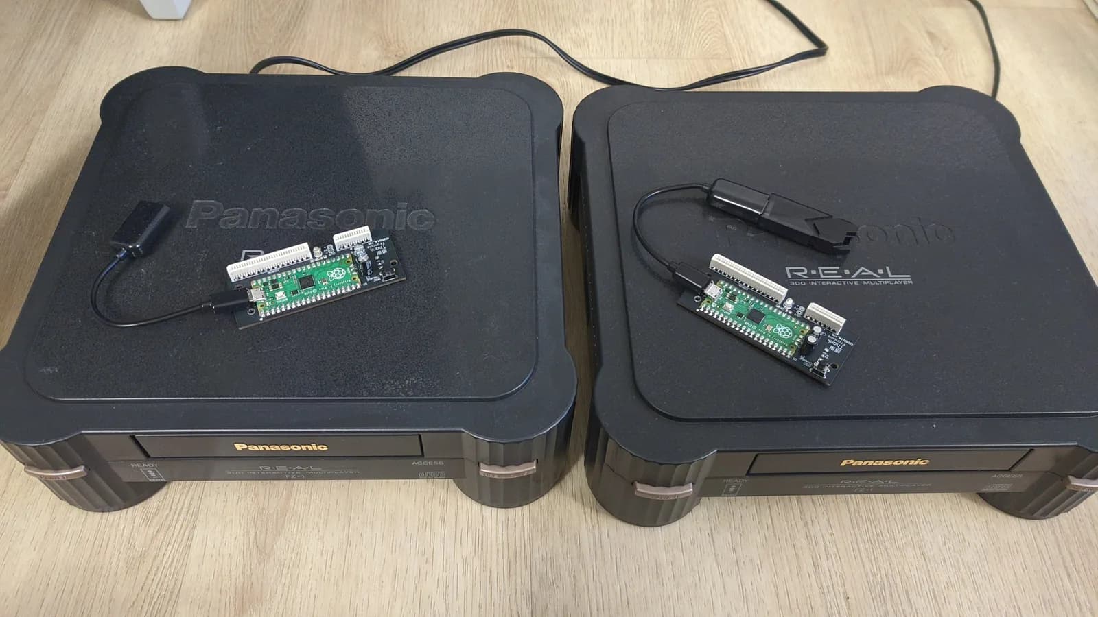 Two closed Panasonic FZ-1 3DO consoles side by side on a wood floor, each with an assembled ODE board (a green Raspberry Pi Pico on a small black board) resting on its lid and wired to a black USB adapter.