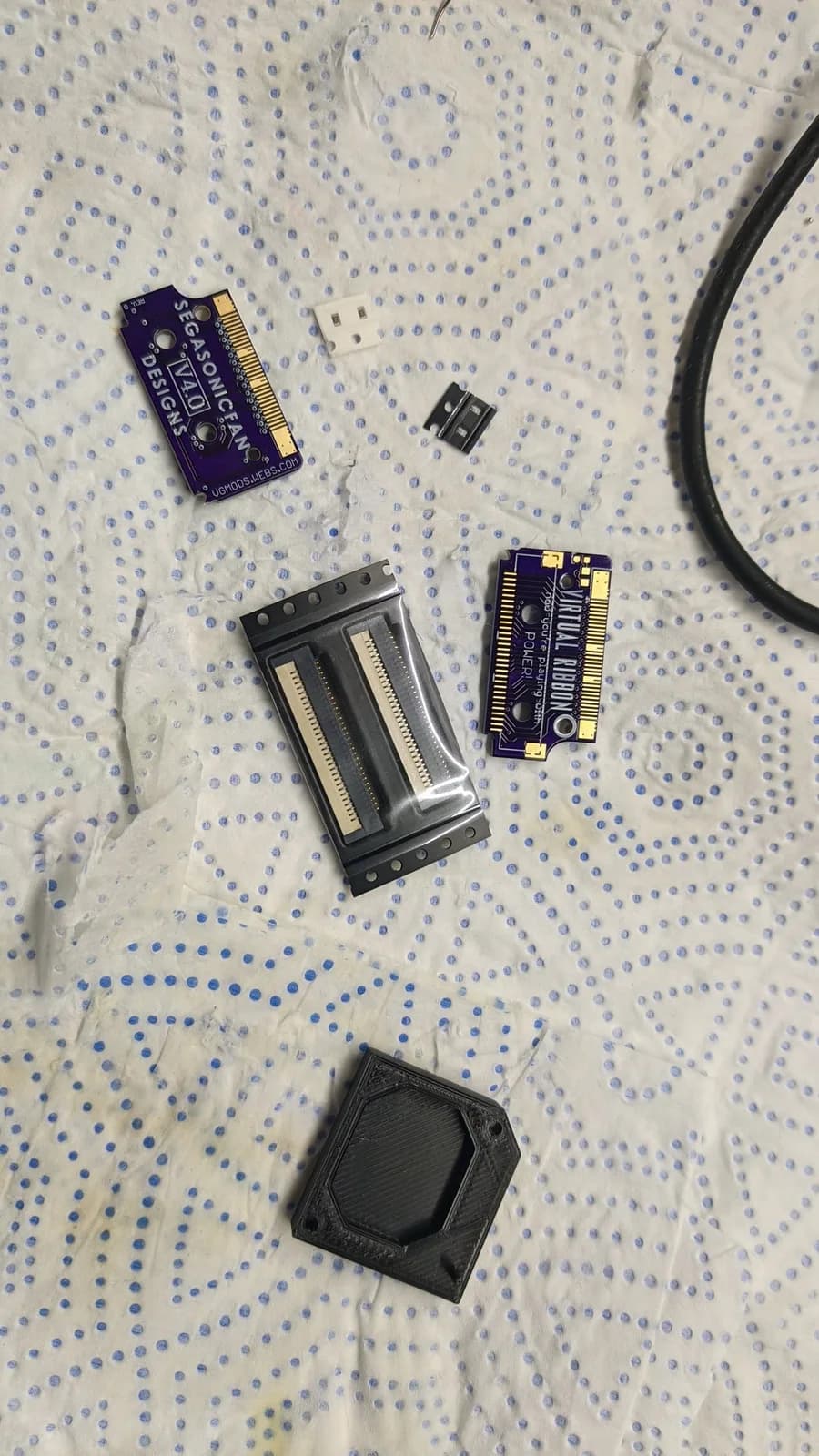 Virtual Boy ribbon cable kit components laid out: purple circuit boards, metal mounting bracket, connector piece, and black plastic housing on dotted fabric.