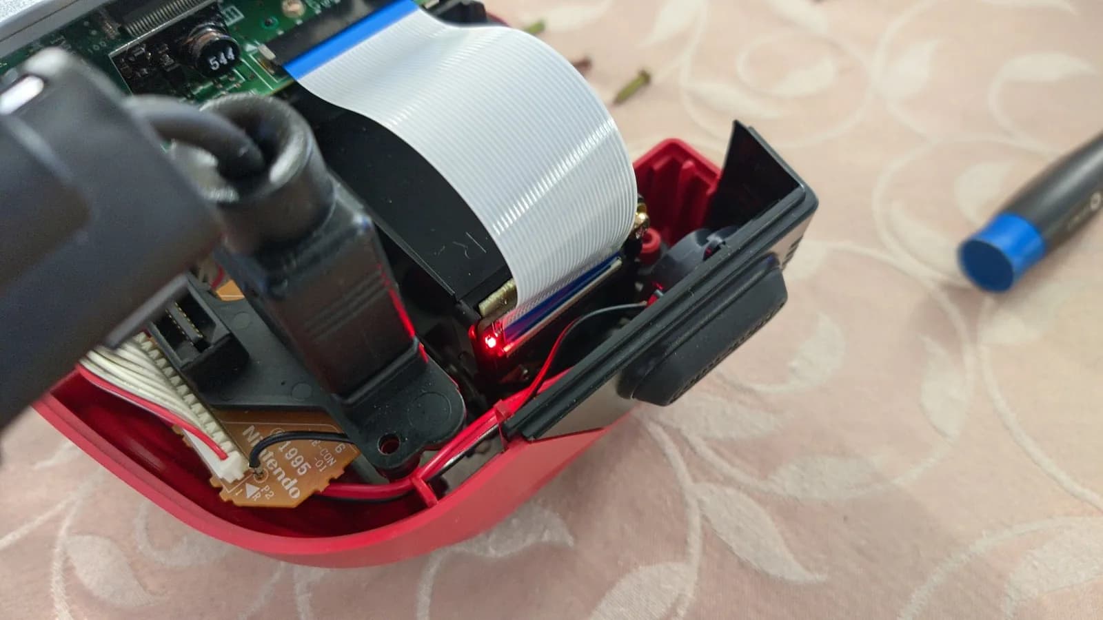 Virtual Boy PCB with white ribbon cable and red mounting bracket during refurbishment; soldering mat visible below.