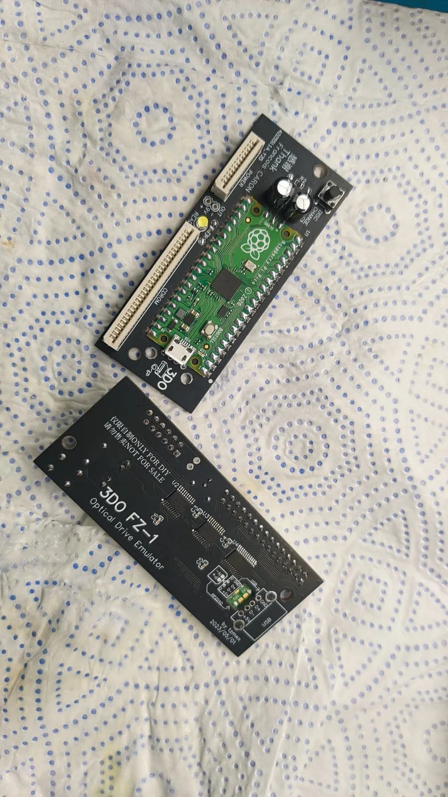 Two assembled 3DO FZ-1 ODE boards on a paper towel: the top board seen from the component side with a green Raspberry Pi Pico, black electrolytic capacitors, a green LED and a button; the second board shown from its underside, its shiny solder joints visible and the back of its Pico showing through the cutout in the black PCB.