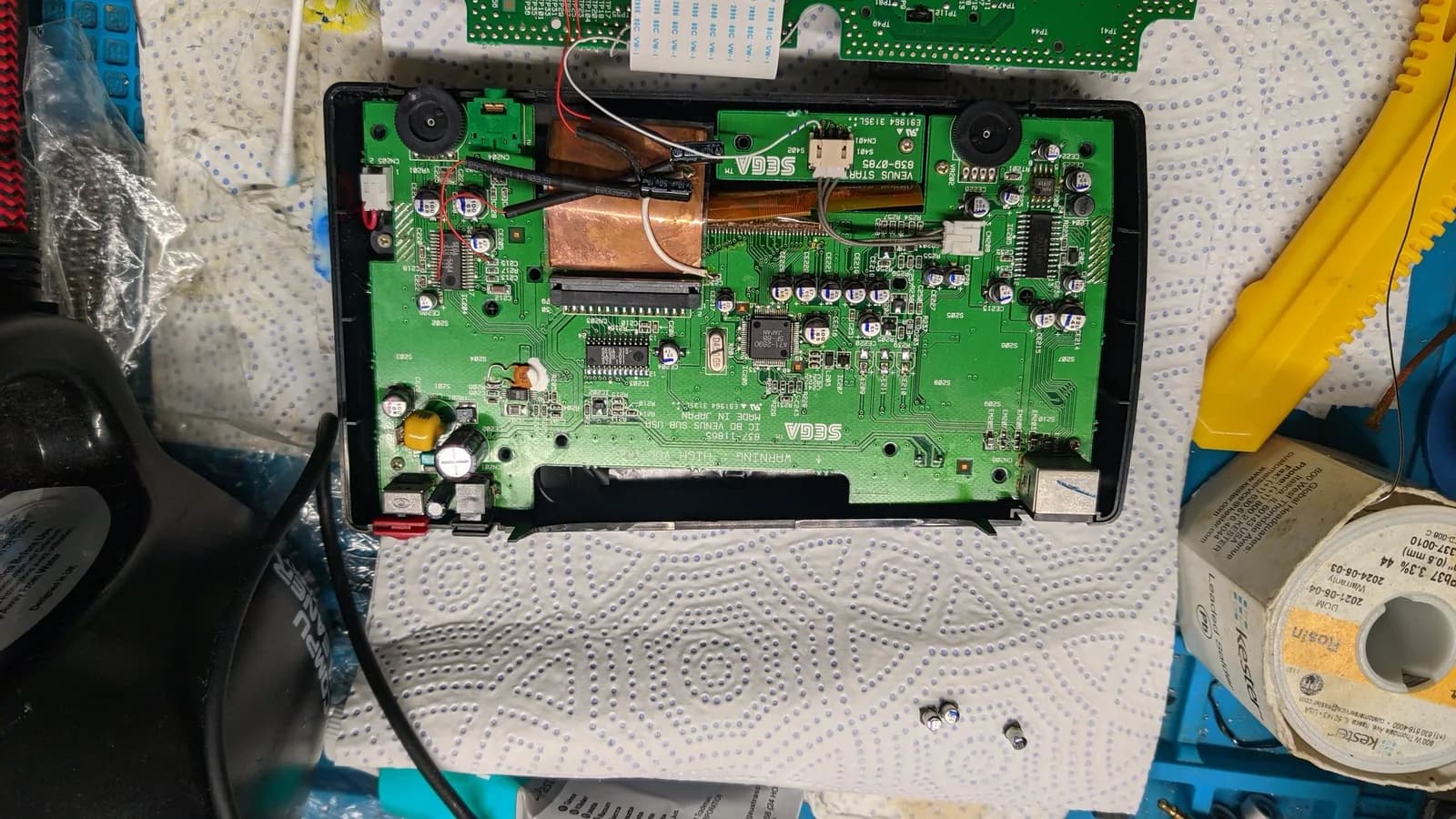 Sega Nomad mainboard undergoing capacitor replacement; green PCB with exposed components, soldering iron, and desoldered capacitors visible during restoration.