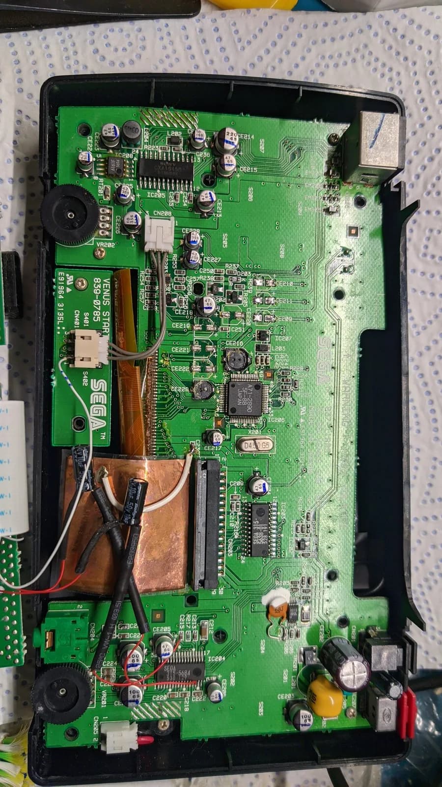 Sega Nomad motherboard with capacitors removed, copper heat sink visible, soldering iron nearby, oxidation and corrosion evident on board surface.