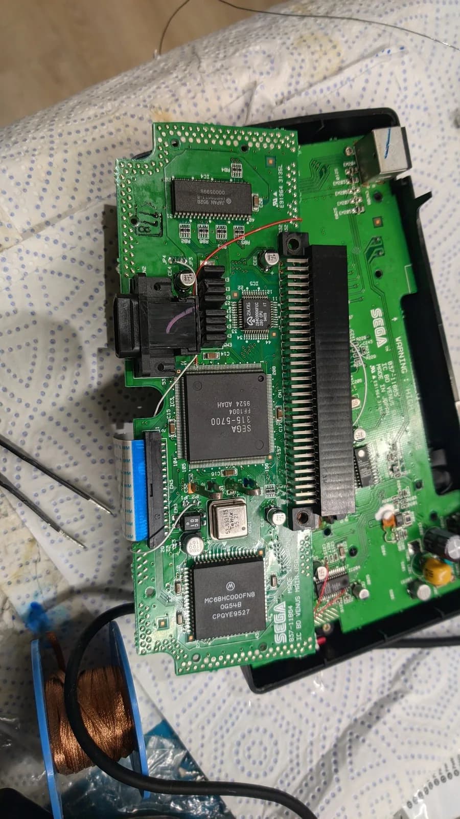 Sega Nomad motherboard with exposed chips and capacitors during restoration; soldering iron and desoldering tool visible on white mat.