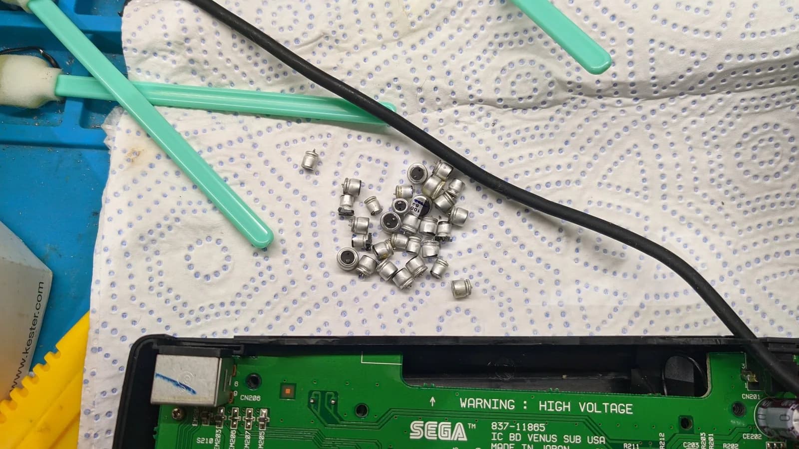 Disassembled electrolytic capacitors scattered on perforated work mat next to green and black tools, Sega circuit board visible below.