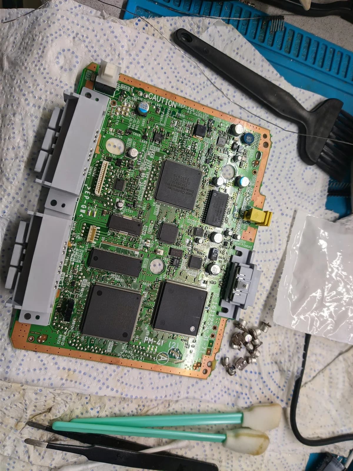 Fully recapped green Sony PSOne mainboard on a paper towel over a work mat, its new capacitors fitted and the old SMD electrolytics that were removed piled in a small heap of silver cans to the right, with tweezers, a spudger and flux-stained cotton swabs around it.