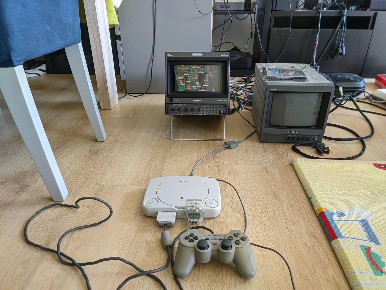 The reassembled white PSOne (SCPH-101) with a grey DualShock controller and a clear PocketStation on a wood floor, running Gran Turismo on a Sony BVM-D9H5, a second smaller Sony monitor beside it with a Gran Turismo game case on top.
