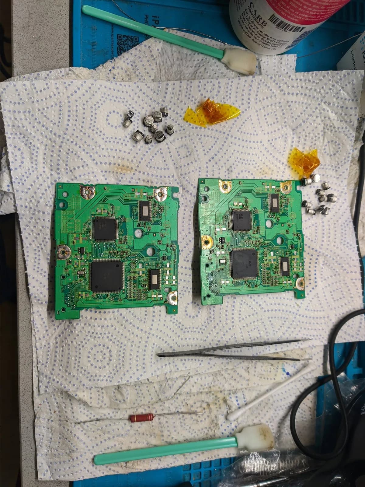 Two GameCube optical drive PCBs being recapped, with desoldered electrolytic capacitors, tweezers, and a resistor laid out on absorbent mat.