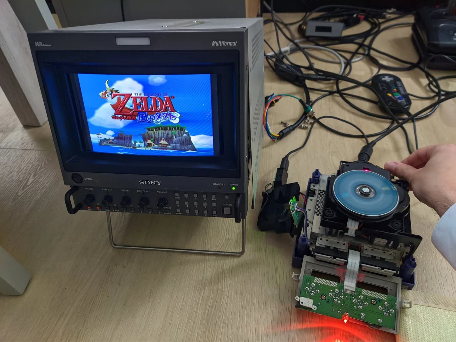 GameCube optical drive internals held open, showing motor and disc mechanism with multimeter probe measuring resistance during diagnostic testing.