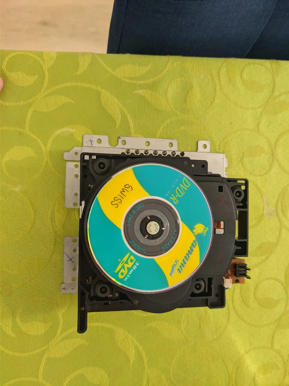 GameCube optical drive with teal and yellow disc label, mounted in black plastic housing with white bracket and flex cable visible.