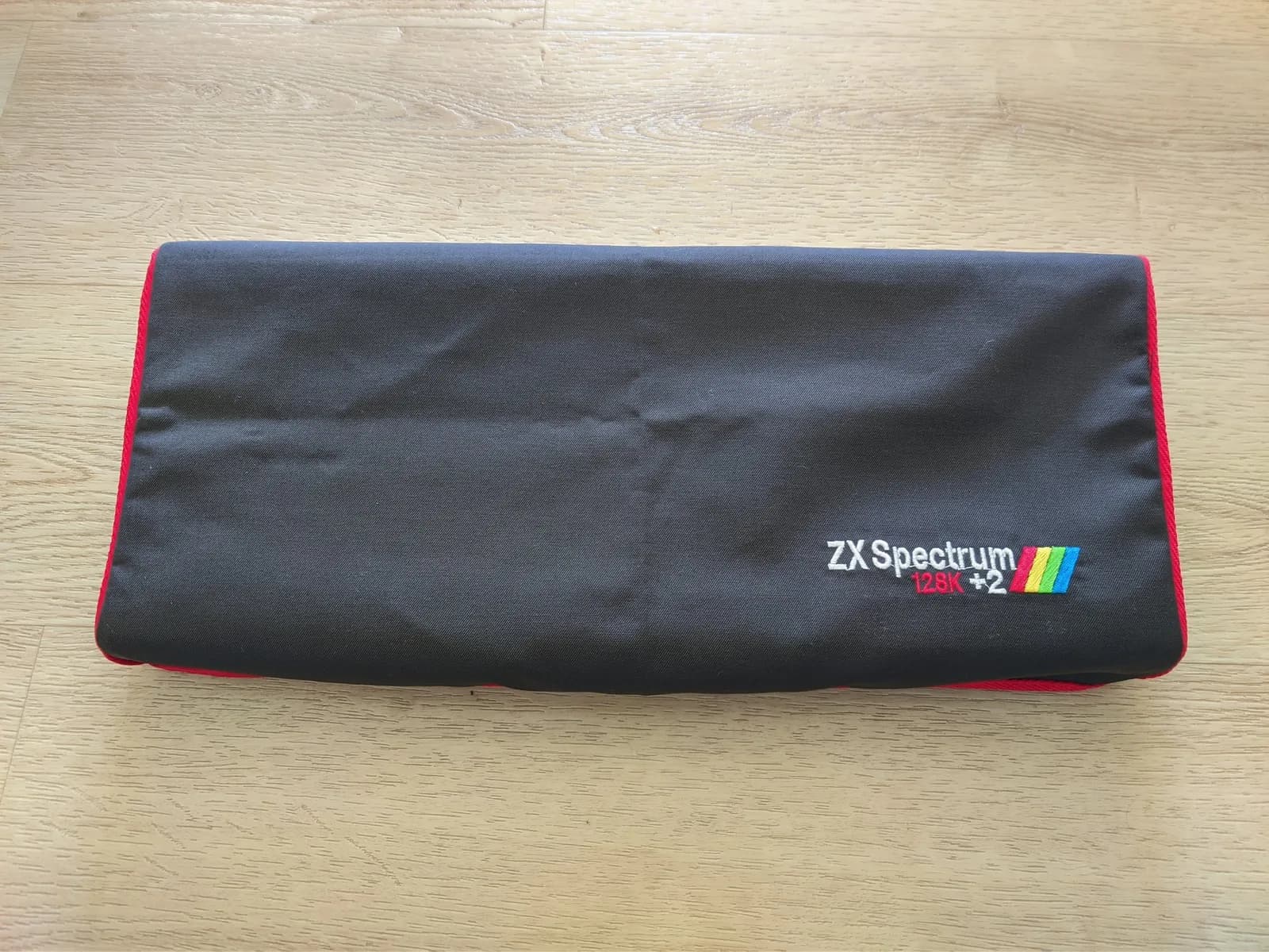 ZX Spectrum +2 dust cover with rainbow logo on black fabric, red trim visible along edge, placed on wooden surface.