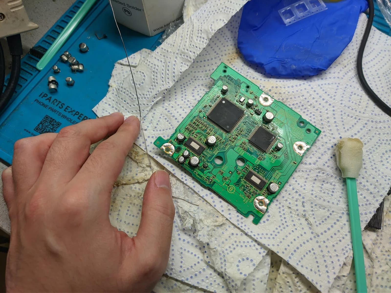 A green GameCube optical-drive PCB with two large black chips, resting on a paper towel on a blue work mat, a hand and a length of solder wire beside it, the removed old capacitors set off to the side and a cleaning swab nearby, during the optical-drive recap.