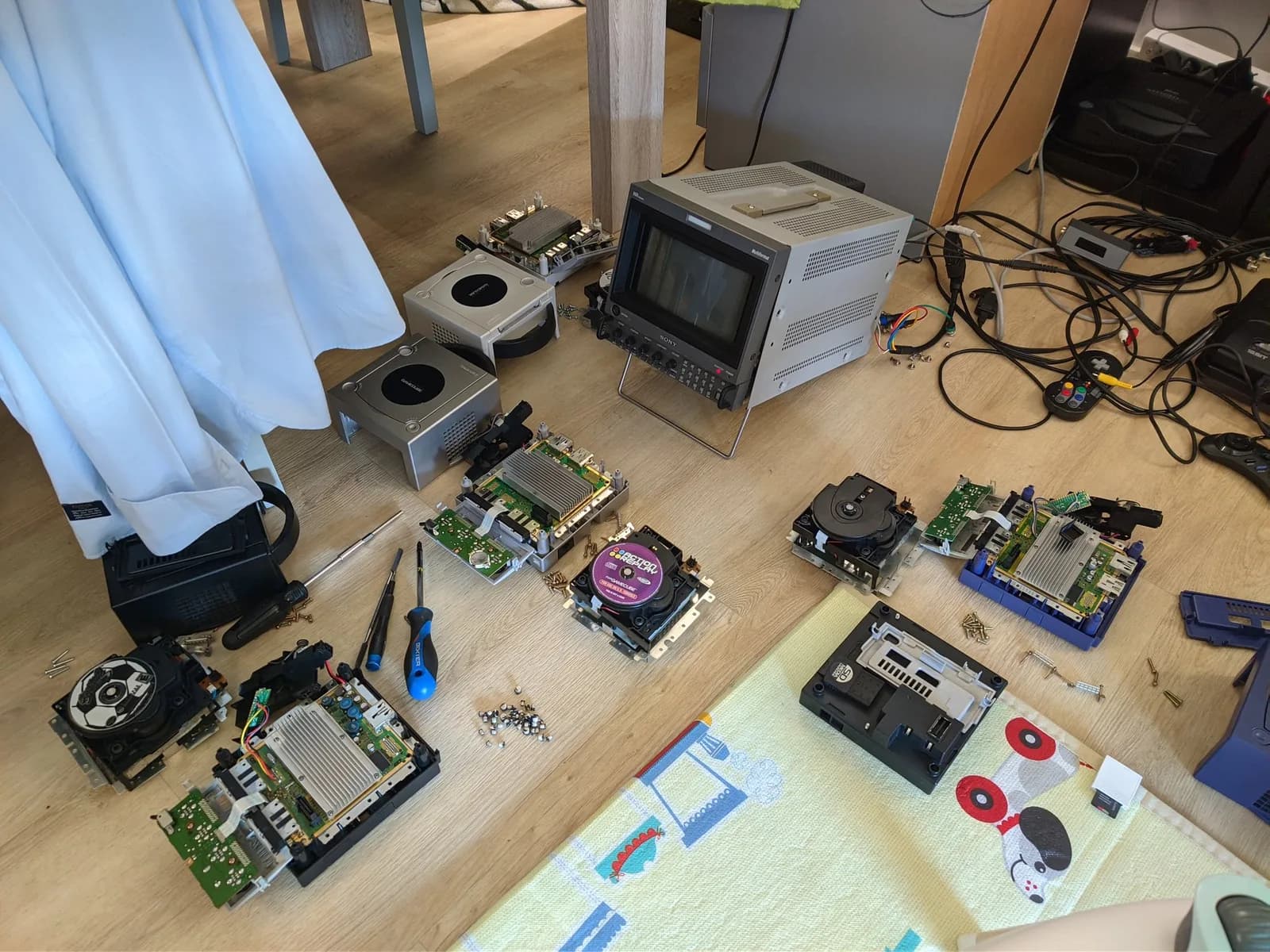 Several GameCube optical drives and mainboards laid out during the fleet maintenance day, with the Sony BVM-D9H5J monitor and tools, an Action Replay disc and an SD Media Launcher, the black GameCube's own drive with Mario Strikers loaded, and a scattering of old electrolytic capacitors on the floor, pulled from the various drives.
