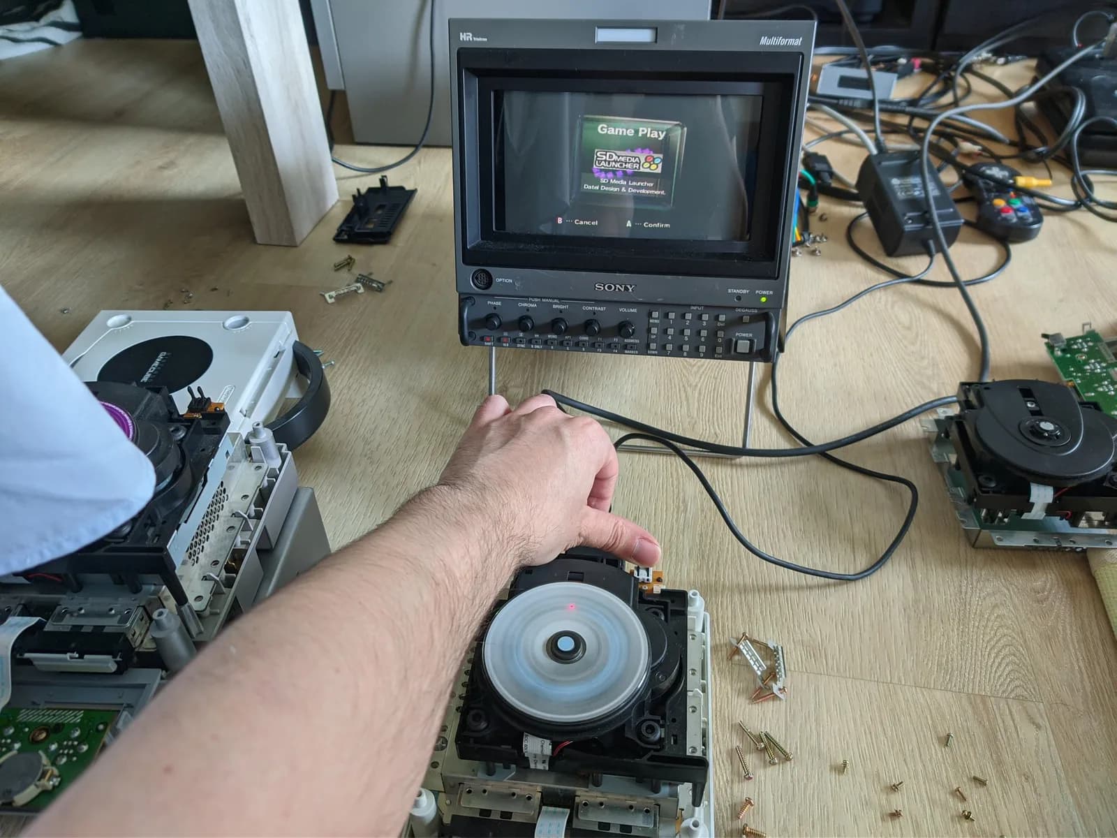 A hand holding a recapped GameCube optical drive, working again after the recap, the Sony BVM-D9H5J monitor showing the GameCube menu's Game Play option.