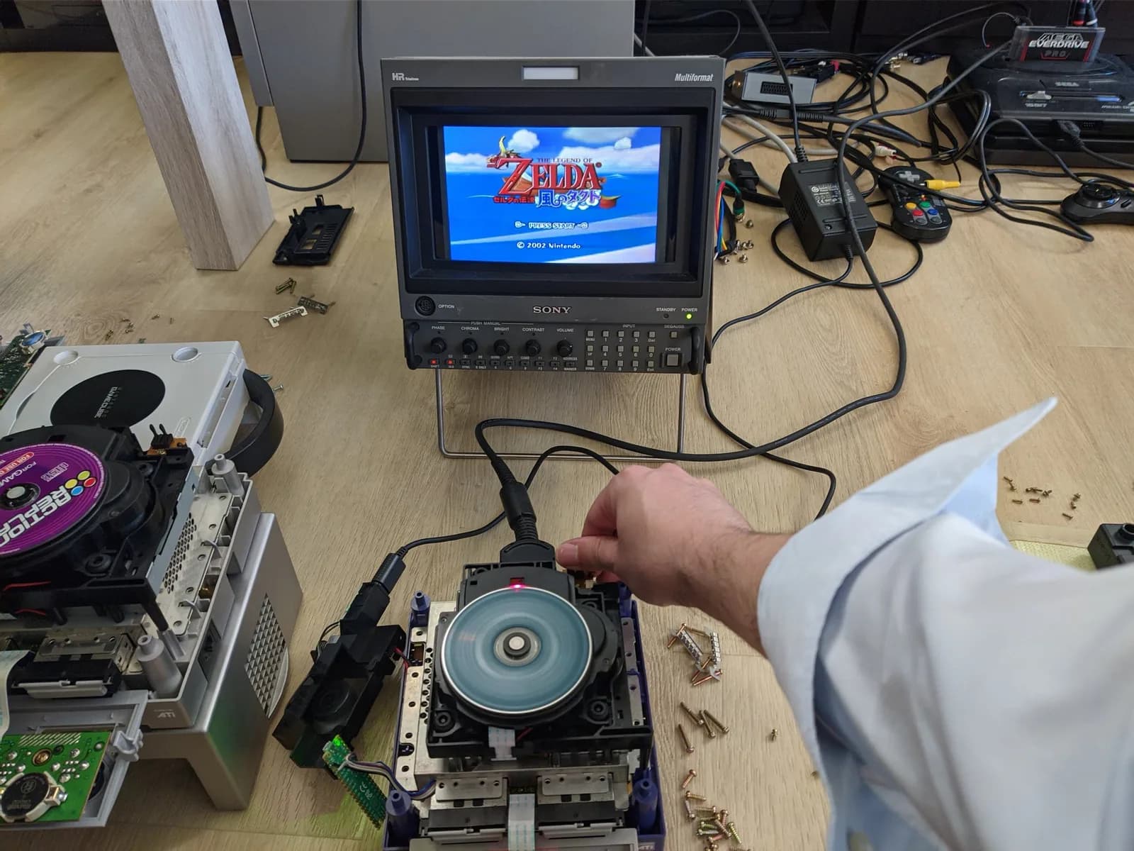 A recapped GameCube optical drive (the indigo unit's) reading a disc on its spindle, the Sony BVM-D9H5J monitor showing the Legend of Zelda: The Wind Waker title screen during testing.
