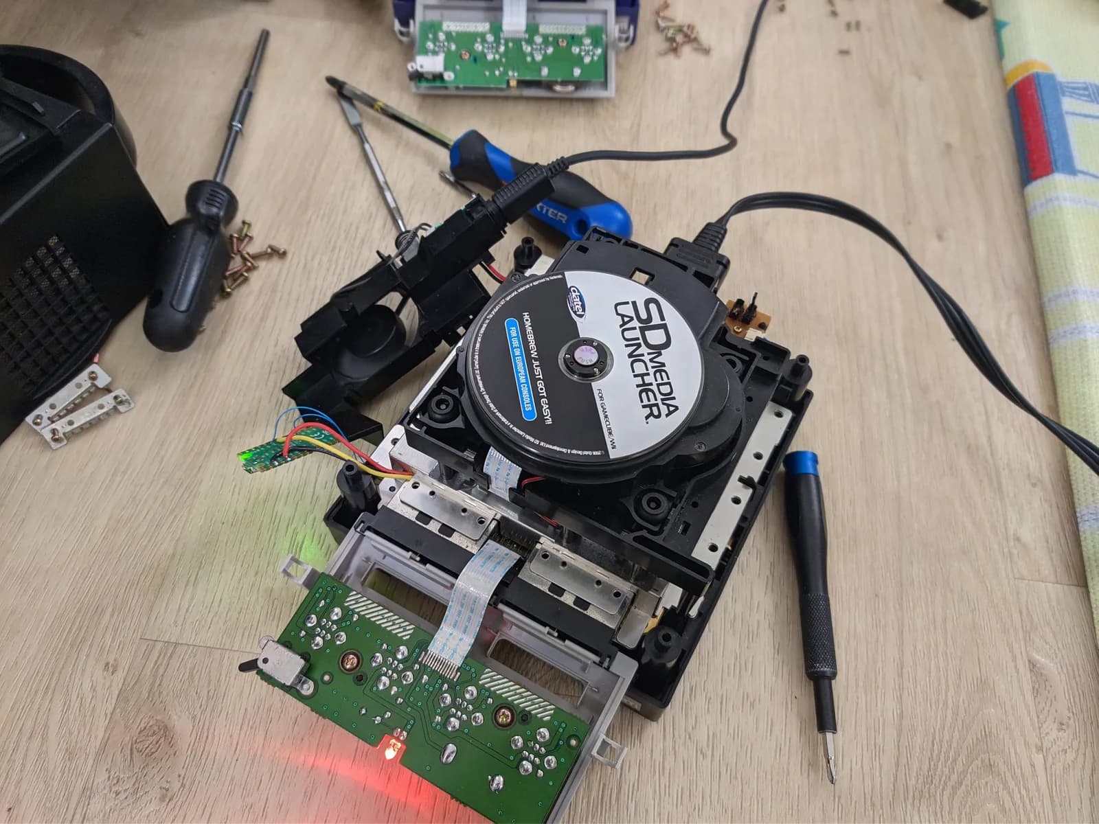 A whole black GameCube connected and powered for testing during the optical-drive recap, its amber power LED lit and its parts out of the shell: the green optical drive with the SD Media Launcher boot disc on its spindle, the controller-port PCB hanging by its flex cable, the cooling fan hanging by the wires of its power-input connector, and the PicoBoot in its 3D-printed holder suspended by its soldered wires, with several screwdrivers alongside on a wood surface.