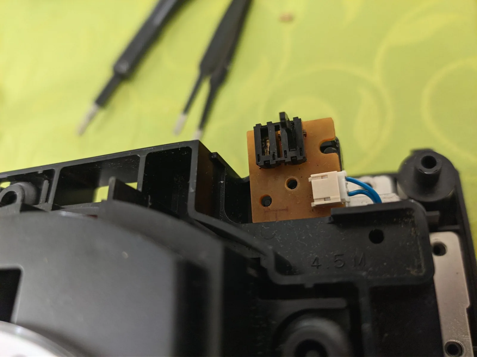 Close-up of the brown topside of the drive's lid-switch board on the black drive chassis, carrying its two through-hole limit switches with one lever snapped off, and a white 2-pin connector with two blue wires, with tools blurred above on a yellow-green surface.