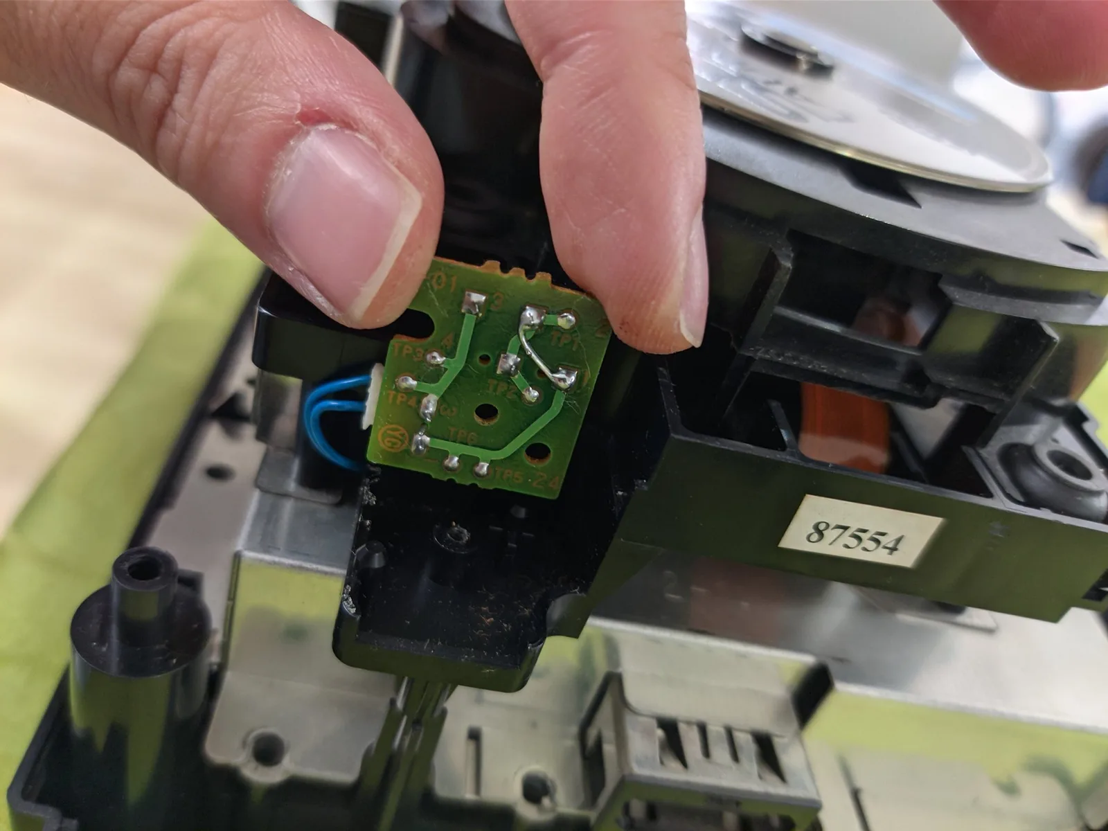 Close-up of the green underside of the same lid-switch board, held in the fingers, where the through-hole limit switches solder to their pads; a component lead has been soldered across to bridge the contacts of the switch whose lever snapped off, so that with that switch permanently jumped closed, the other, intact switch, actuated by the closing lid, lets the console detect the lid-closed signal and start reading discs.