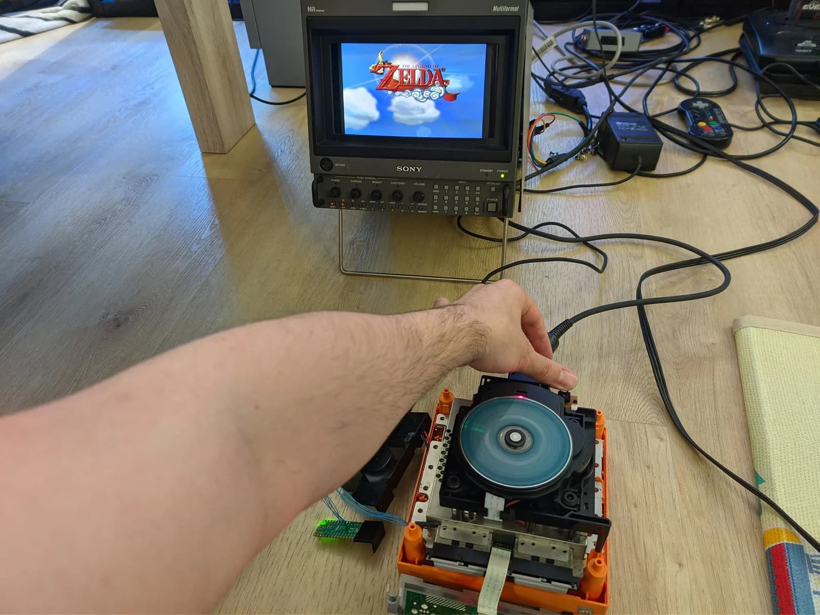Open GameCube optical drive on a wooden floor showing the exposed blue disc and orange housing, with a hand pressing the lid switches that tell the console the lid is closed, testing the drive after recapping its PCB. A Sony BVM-D9H5J monitor behind shows The Legend of Zelda: The Wind Waker.