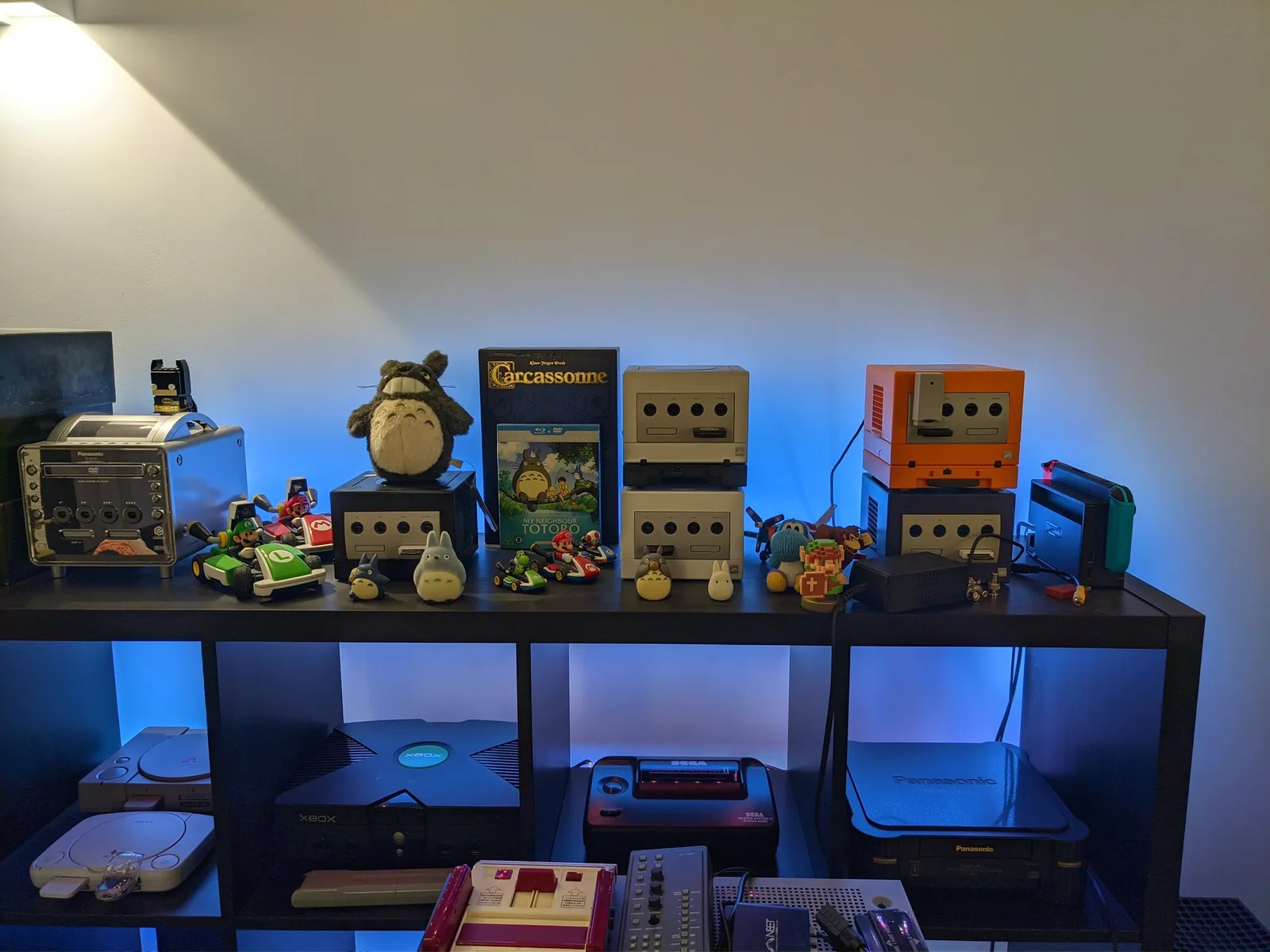 A console collection on a black shelf under blue LED lighting. Top: the silver Panasonic Q, a black GameCube, a silver GameCube, a pearl-white GameCube and a spice-orange GameCube, a docked Nintendo Switch, plus Totoro plushes and Mario Kart figures. Bottom: two PlayStations (a PS1 and a PSone), an original Xbox, a Sega Master System II and a Panasonic 3DO.