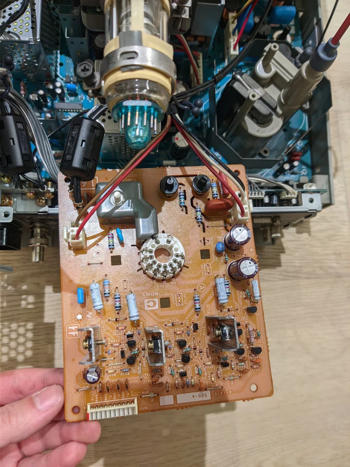 Neck board PCB from Sony PVM-14L2 held in hand, showing capacitors, resistors, potentiometer, and wiring during recap maintenance.