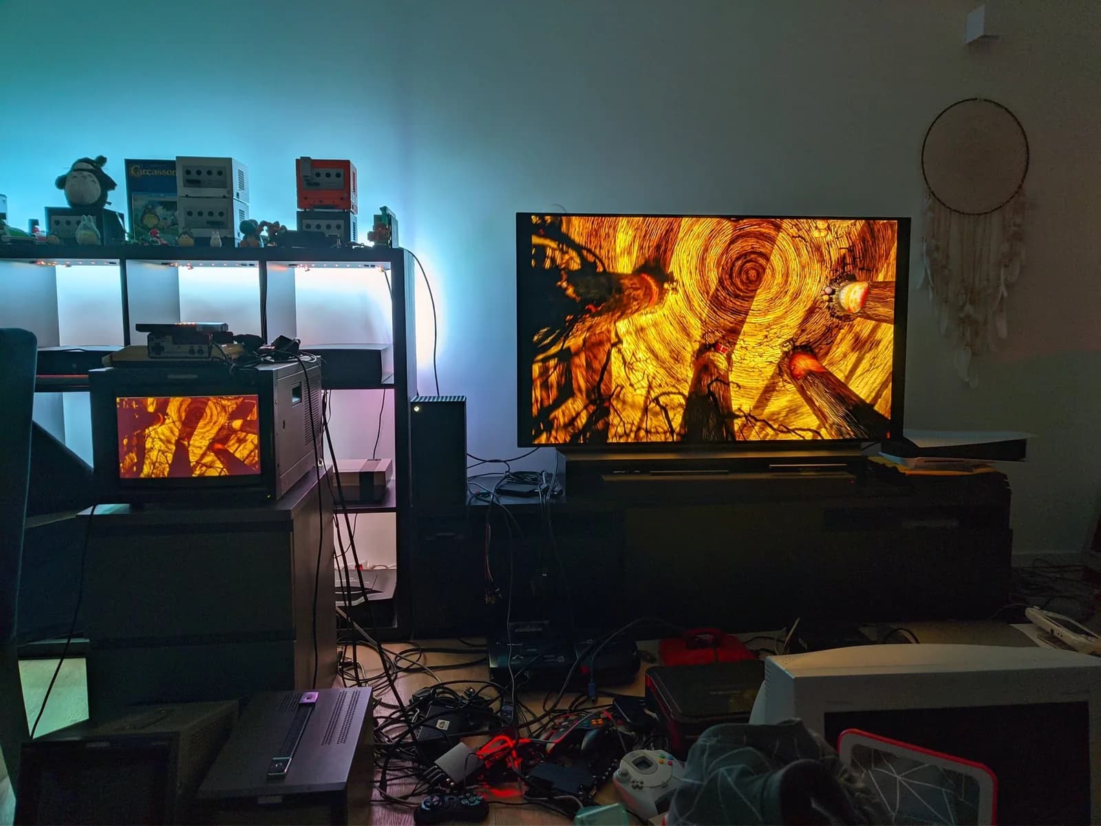 A dark retro-console room, a Sony BVM-D14H1E CRT monitor on the left and an LG C1 OLED TV on the right both showing the opening scene of The Nightmare Before Christmas (a warm orange, spiralling image); GameCube consoles and plush toys on the shelves with LED light strips, cables and controllers across the floor.