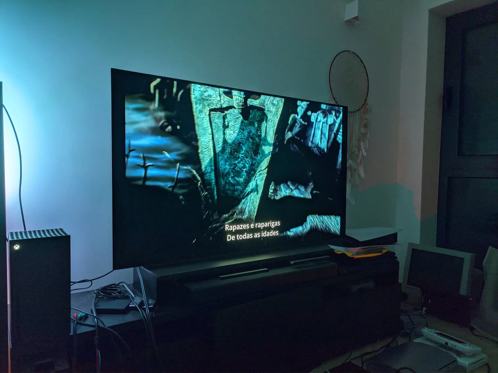 An LG C1 OLED TV showing The Nightmare Before Christmas with Portuguese subtitles, a black Xbox Series X on top of the TV stand, other consoles, a small CRT and cables around the dim room.
