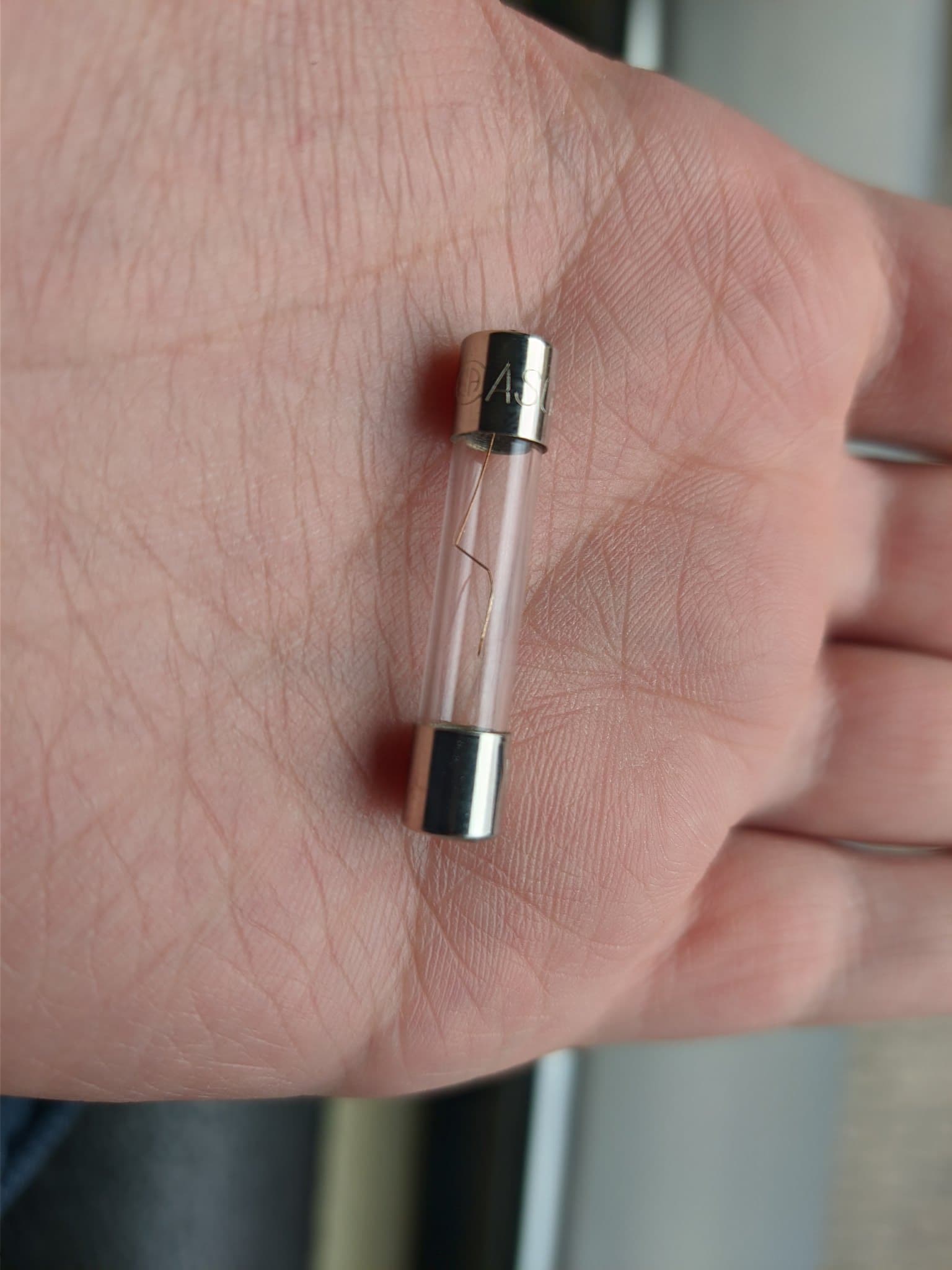 Glass cartridge fuse held in palm, its internal element visibly broken inside the clear glass tube (the fuse failed from mechanical shock).