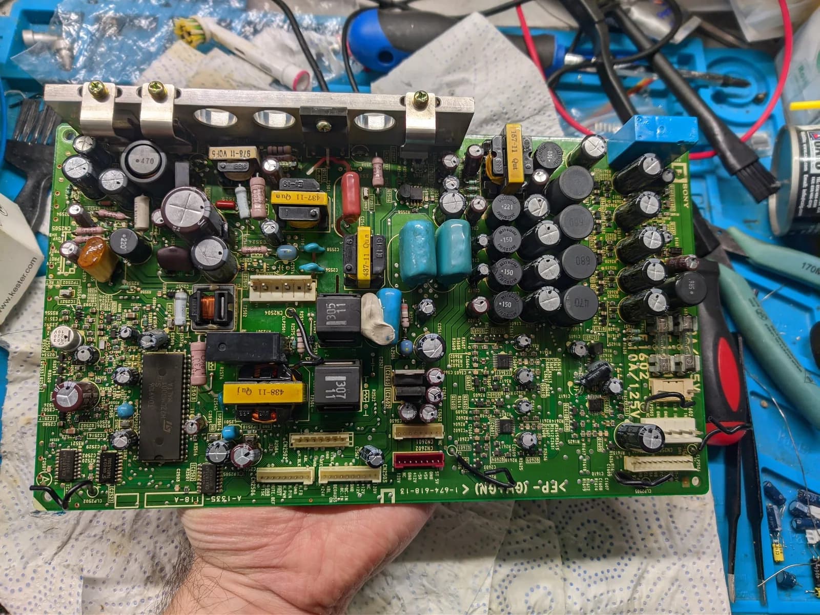 Component side of the Sony BVM-D9H5J D board (deflection board), held in hand: rows of black electrolytic and blue capacitors, ICs and resistors, and a metal heatsink bar along the top, over a workbench mat with tools.