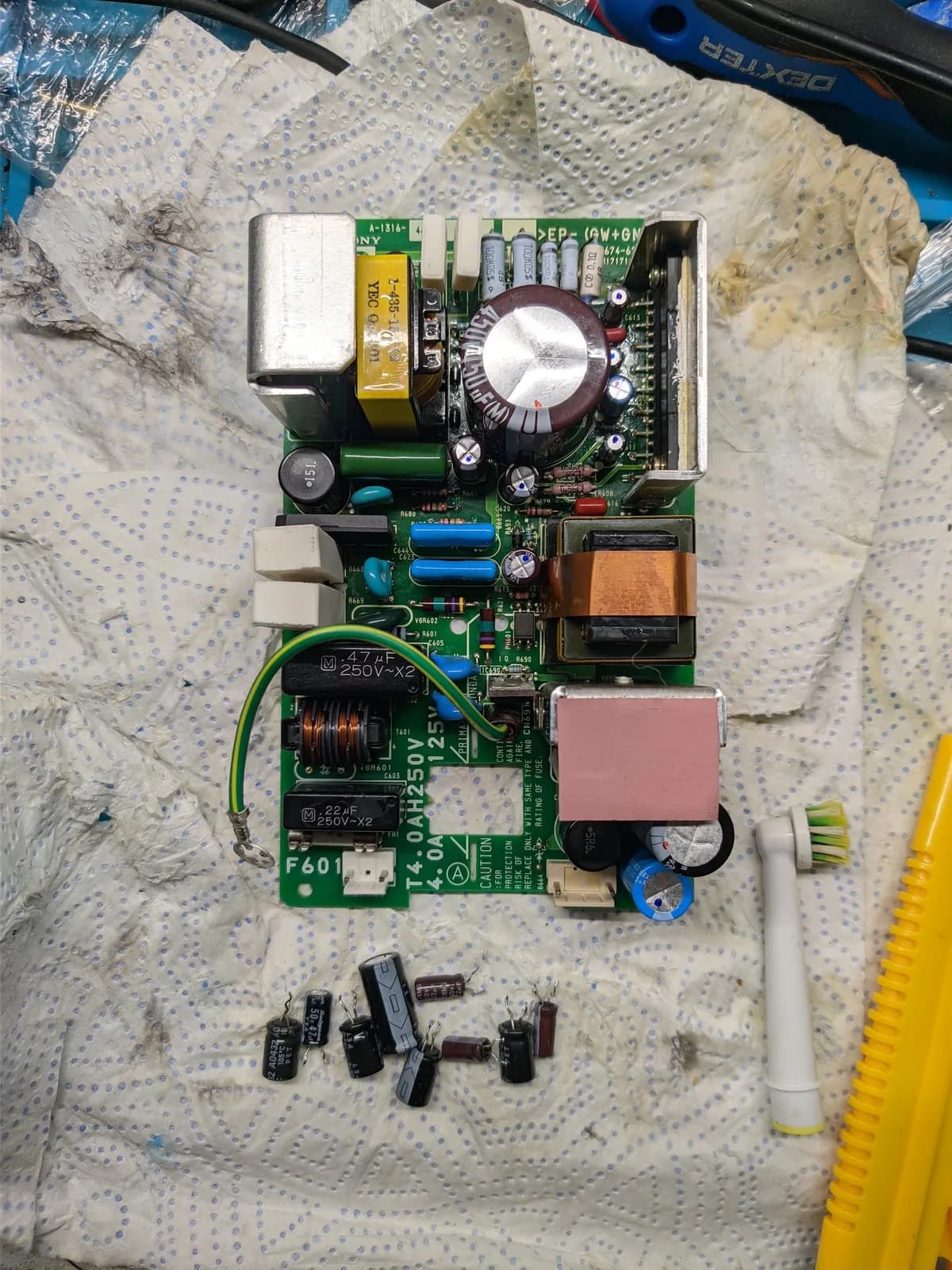 BVM-D9H5J PSU board (component side up) on a paper towel after a partial recap: the large transformer, the large black main filter capacitor (the only cap left in place, since a replacement in that spec would not fit the case), a copper-wound inductor and a pink-insulated component, with the removed old electrolytic capacitors laid out on the towel below.