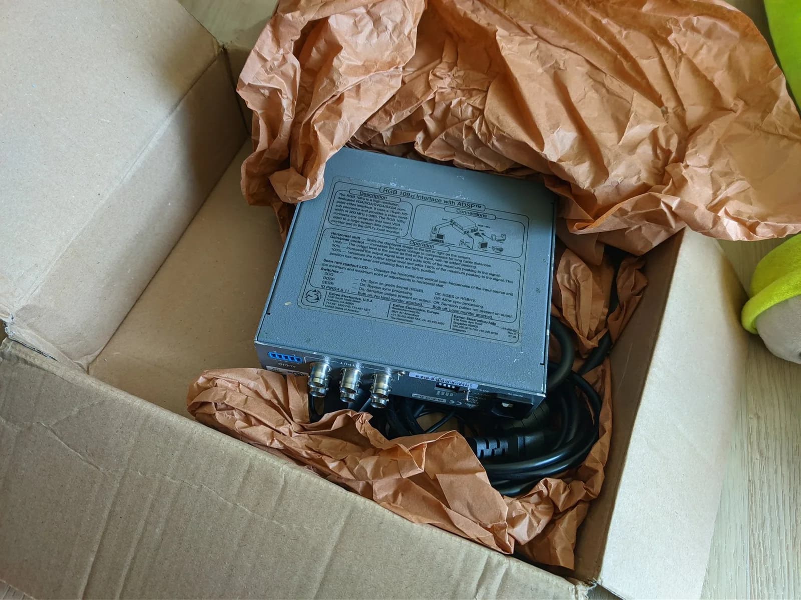 Extron RGB 109xi interface (top label reads 'RGB 109xi Interface with ADSP') nestled in brown kraft paper inside a cardboard box, a dark grey metal unit with six BNC connectors in two stacked rows of three and a coiled black power cable.