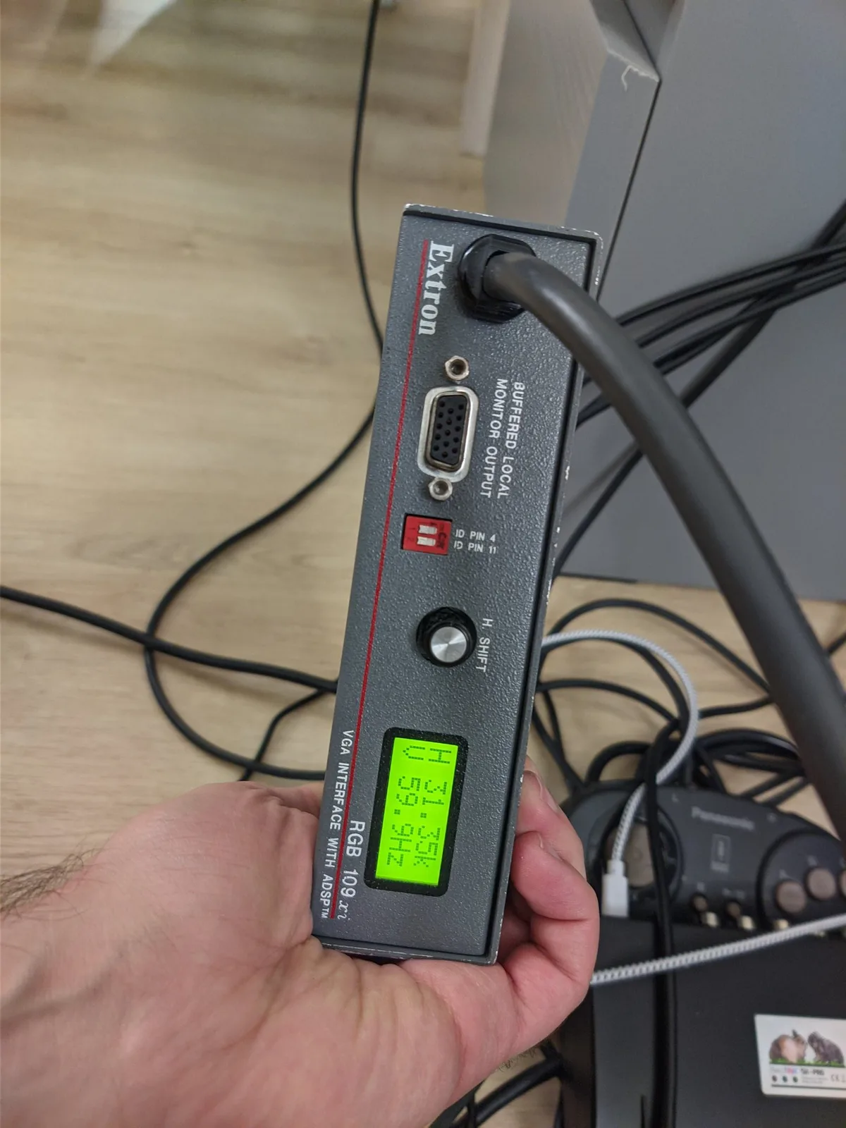 Extron RGB 109xi (VGA Interface with ADSP) held in hand, its green backlit LCD showing a scan-rate readout of 31.35 kHz and 59.9 Hz, with the VGA buffered local monitor output connector and a red ID-PIN DIP switch on the front panel and cables on the floor behind.