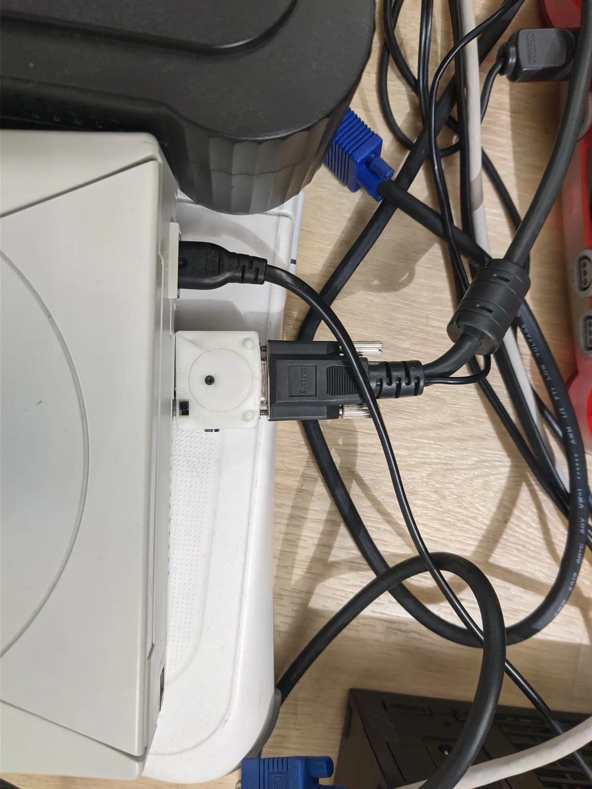 Sega Dreamcast fitted with Jeff Chen's DC2VGA dongle on a wood surface, its figure-8 power lead plugged in, a black Extron-labelled VGA connector mated to a white adapter block and a blue VGA plug among tangled black cables; this is the Dreamcast that was outputting to the Extron device.