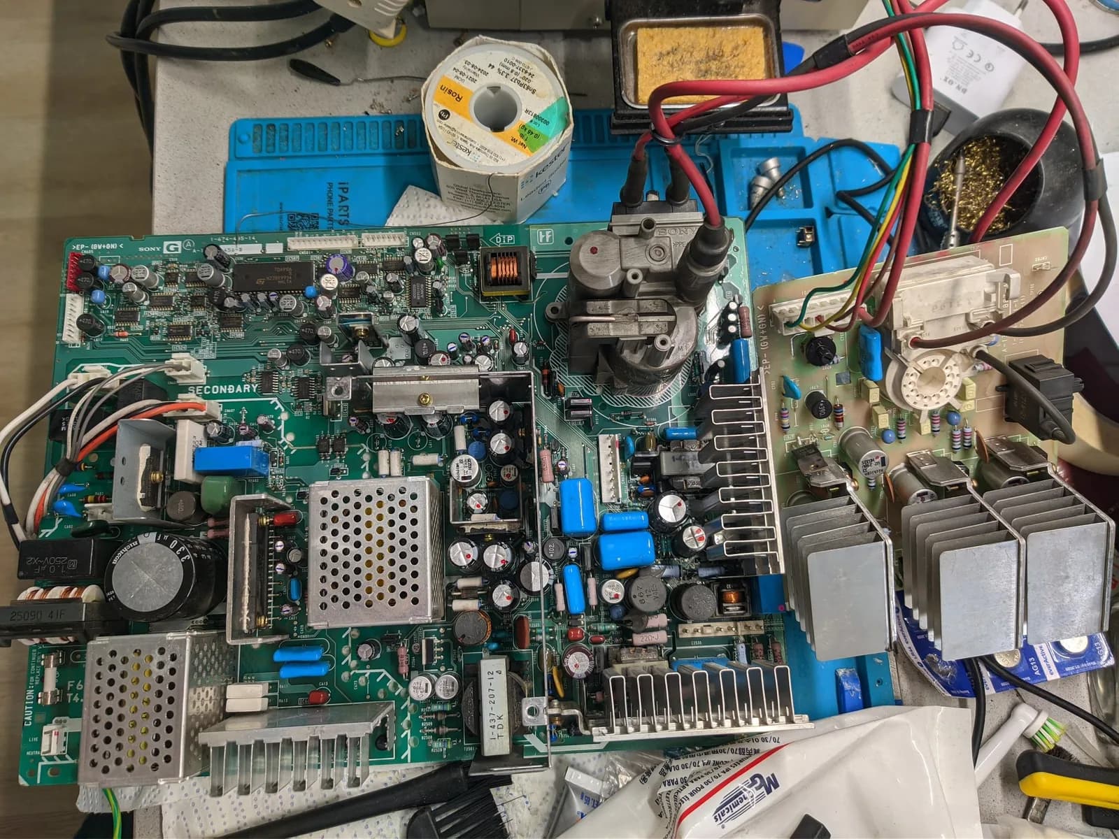 The Sony BVM-D14H1E's G board and neck board out on a blue silicone work mat during servicing, with a cream Kester leaded-solder spool (SN63PB37), a brass-wool tip cleaner, a sponge, and red and black wires around them.