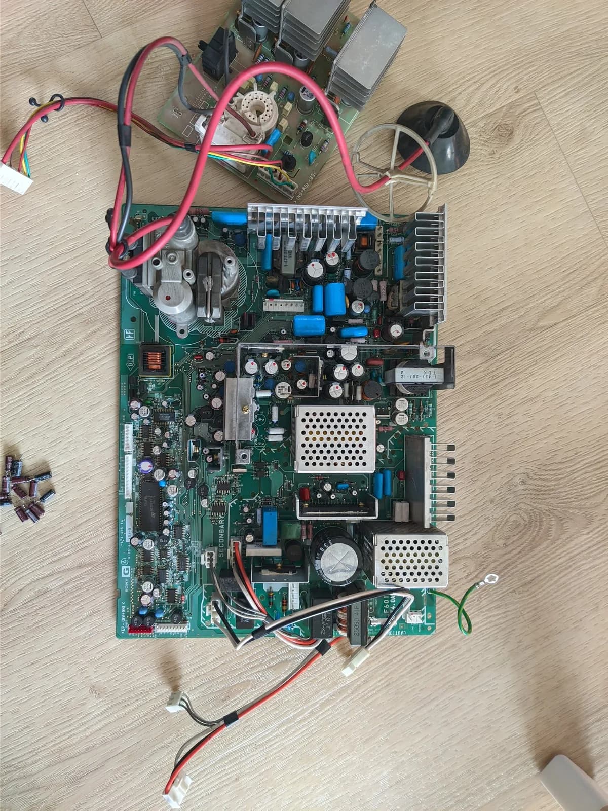 The Sony BVM-D14H1E's G board and neck board on a wood floor after recapping, the new caps fitted and the removed old electrolytics (brown and black) lying beside them, with finned heatsinks and red and black cables linking the two boards.
