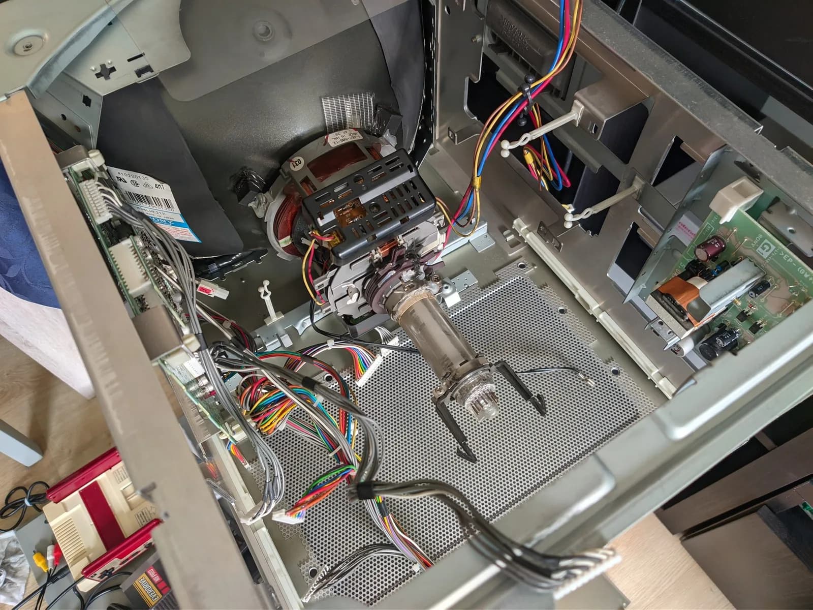 The Sony BVM-D14H1E almost completely disassembled during the recap, its boards removed, exposing the CRT rear, deflection yoke and glass tube neck on the perforated metal chassis with the wiring harnesses; a Nintendo Famicom with an EverDrive N8 Pro box sits on top of another monitor on the floor alongside.
