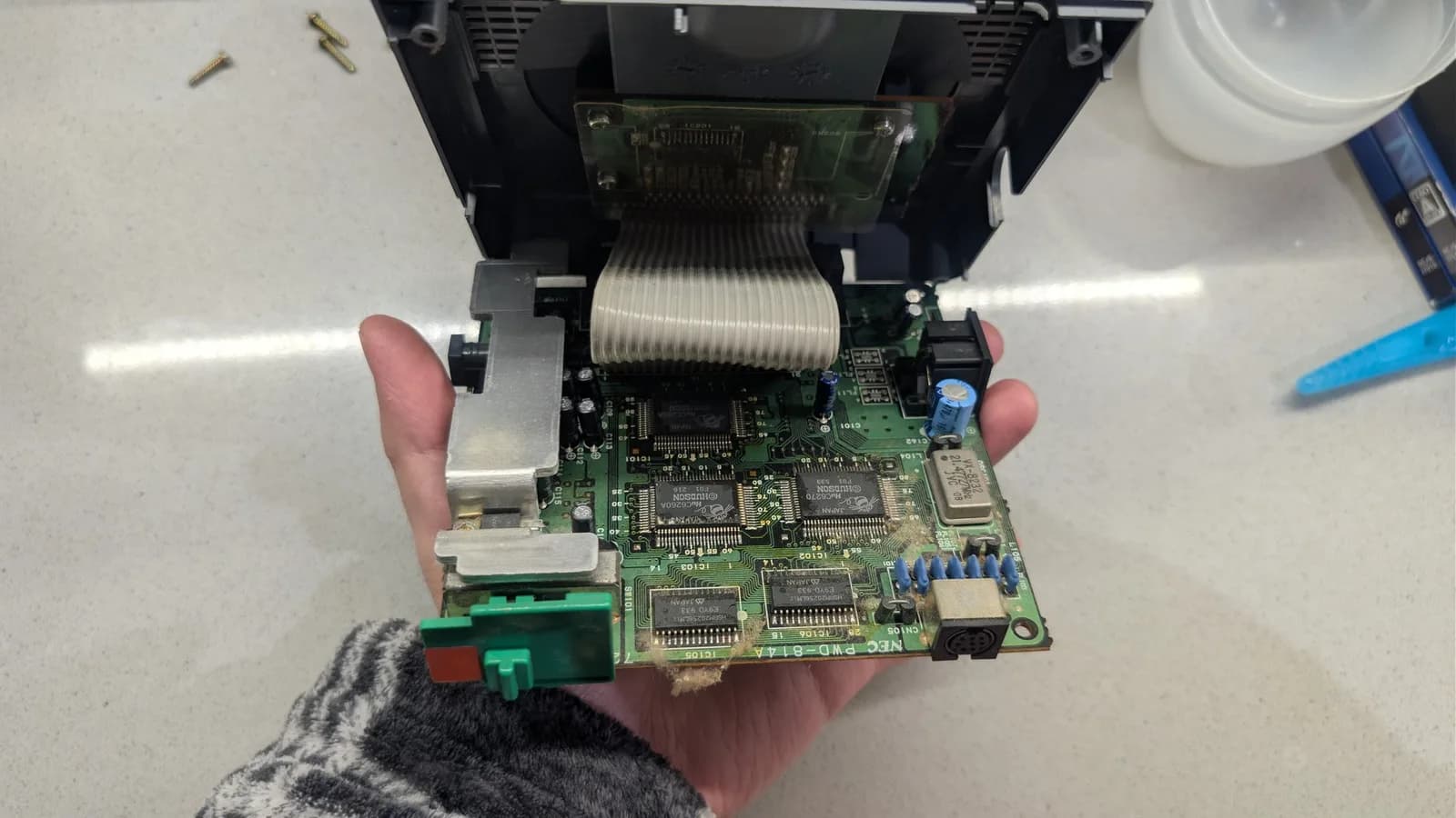 NEC PC Engine CoreGrafx mainboard before the recap, held in hand, dust and fluff over the board, with the ICs, voltage regulator (under its metal heatsink bracket) and the metal-can crystal oscillator visible.
