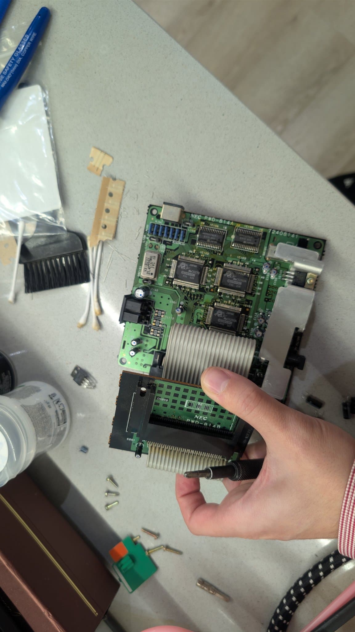 Hand holding the recapped NEC PC Engine CoreGrafx mainboard along with a screwdriver, the new capacitors, voltage regulator and ICs visible.