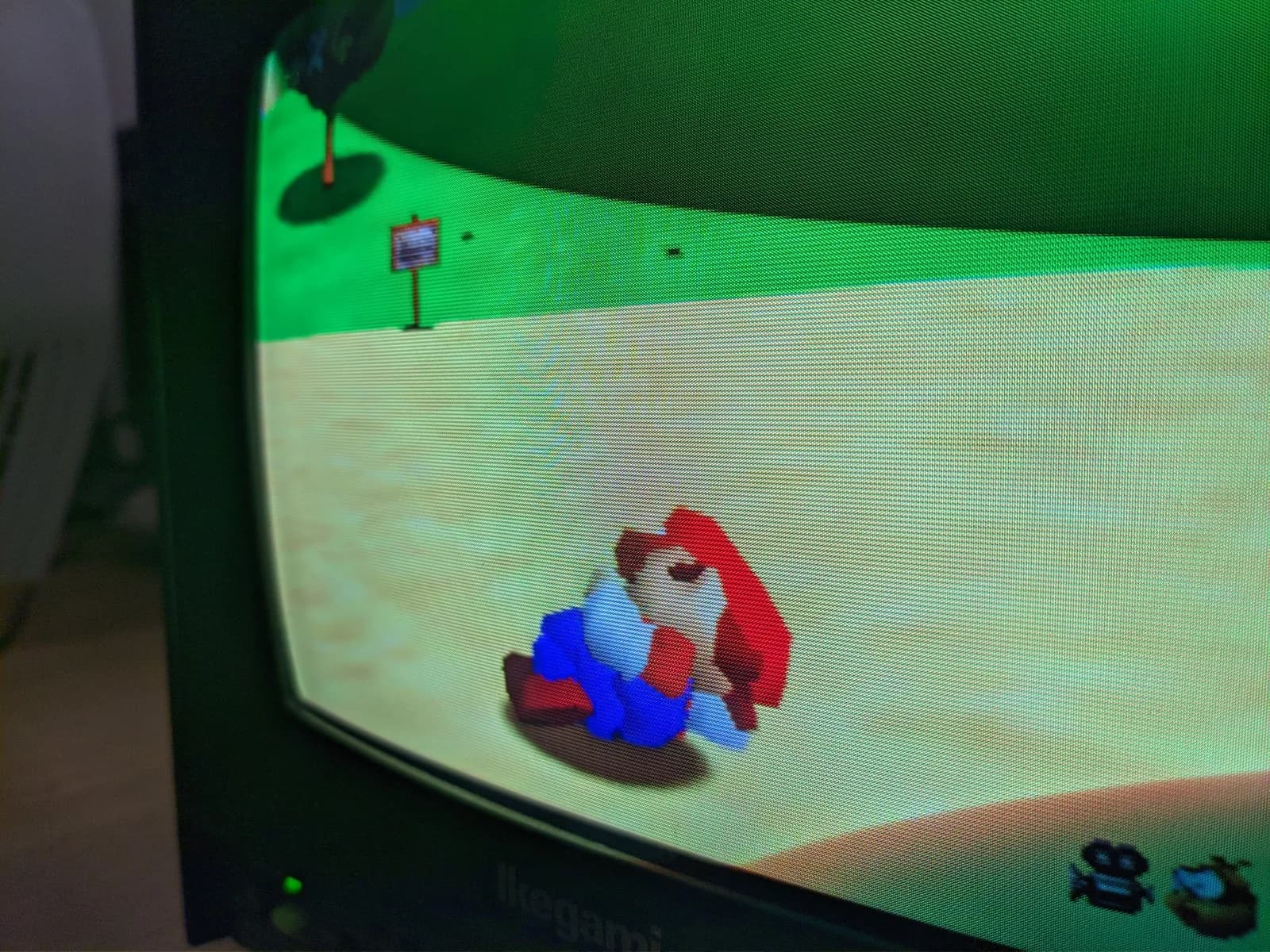 Super Mario 64 on the Ikegami CRT (Ikegami badge visible at the bottom), Mario in his idle, sleeping pose lying on the sandy ground outside the castle.