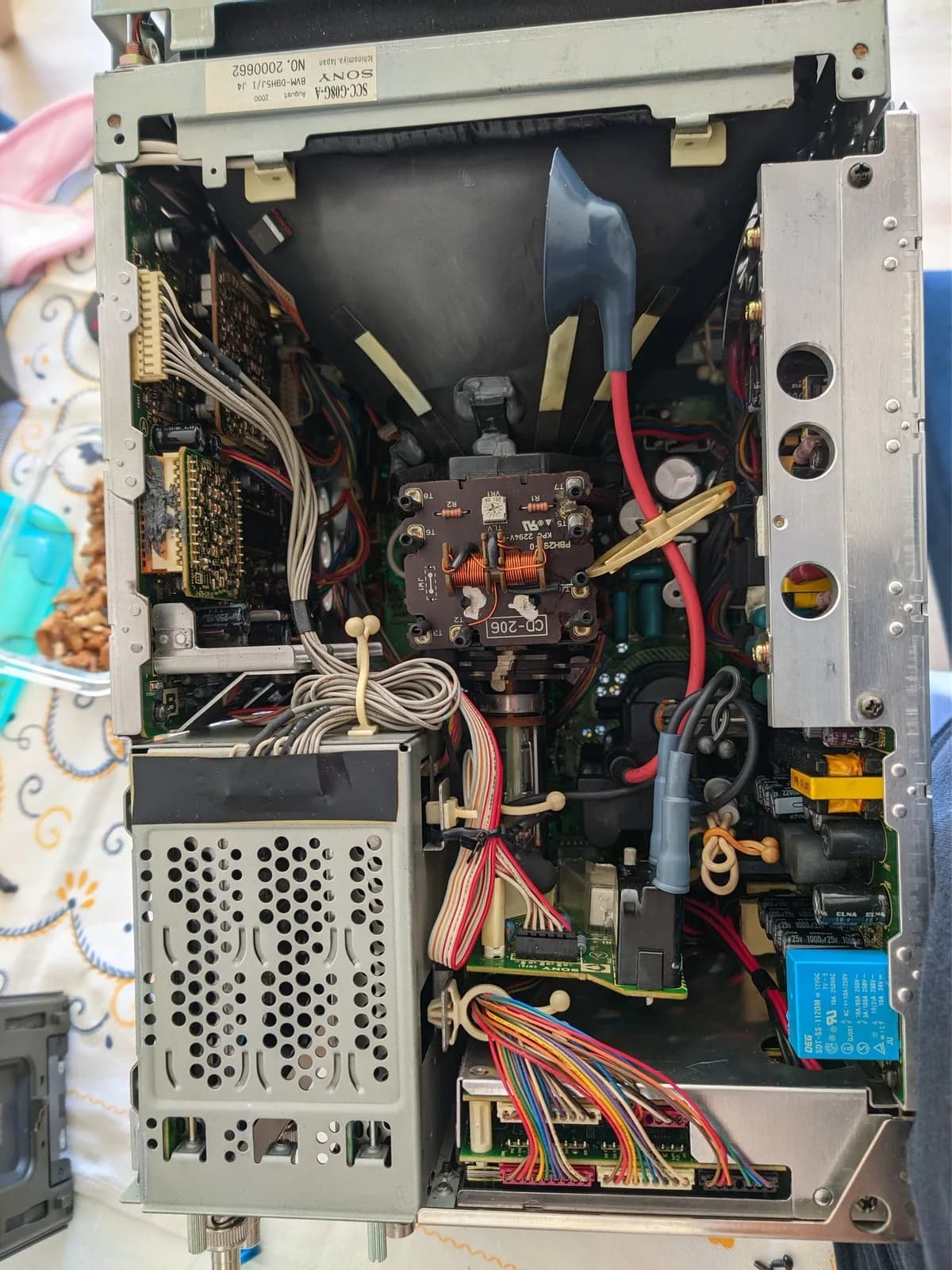 Interior of the opened Sony BVM-D9H5J during servicing: the CRT neck and deflection yoke at centre with the anode cap detached from the tube, the black perforated PSU module at lower left, and multiple PCBs with multicoloured wiring harnesses (a blue relay on the right board), the interior looking clean.