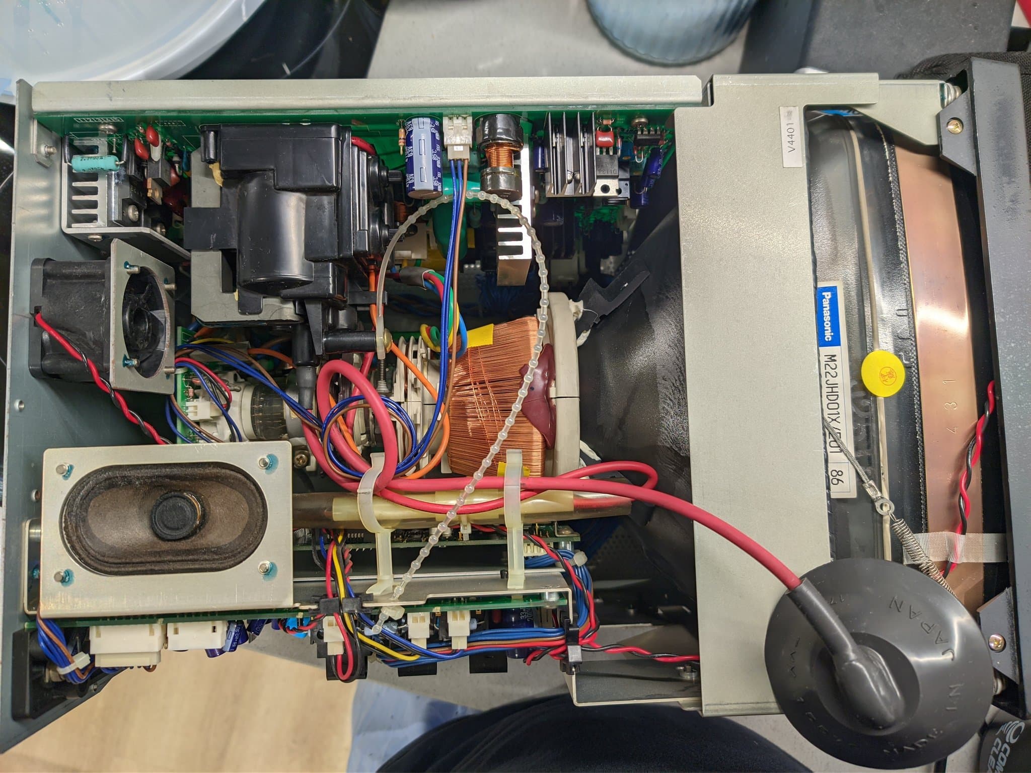 Open Ikegami TM10-17RA interior during the cleaning pass, from another angle: a speaker, a copper coil and transformer, heatsinks, a Panasonic-labelled component, and red and blue wiring.