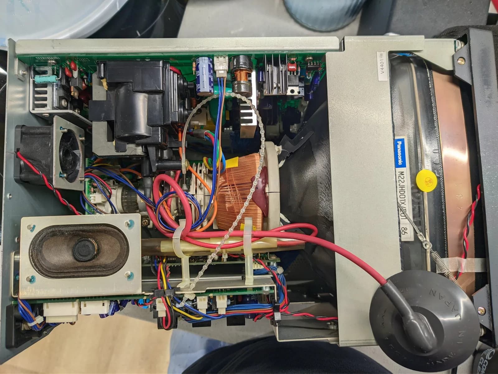 Open Ikegami TM10-17RA interior during the cleaning pass, from another angle: a speaker, a copper coil and transformer, heatsinks, a Panasonic-labelled component, and red and blue wiring.