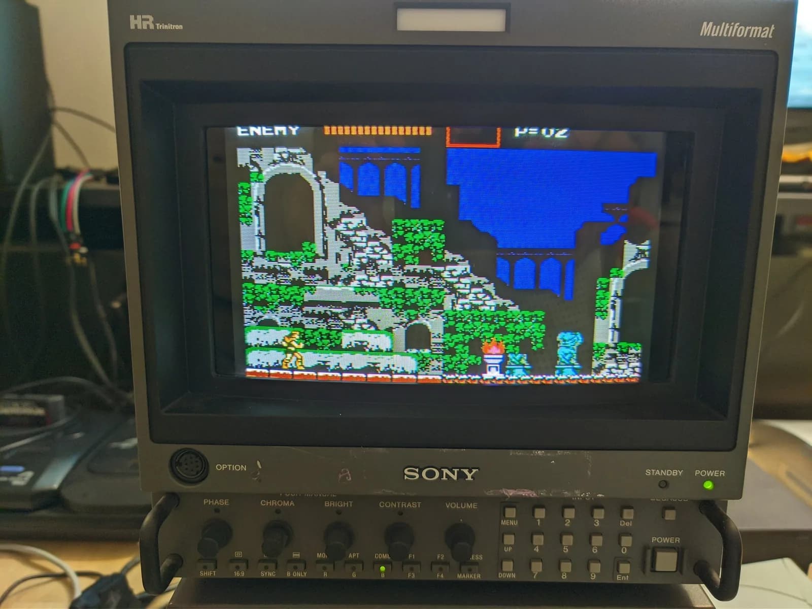 Sony BVM-D9H5J broadcast CRT (HR Trinitron, Multiformat) displaying Castlevania III: Dracula's Curse, Trevor Belmont wielding a whip in a ruined stage with green foliage and a blue background, power LED lit, on a desk.