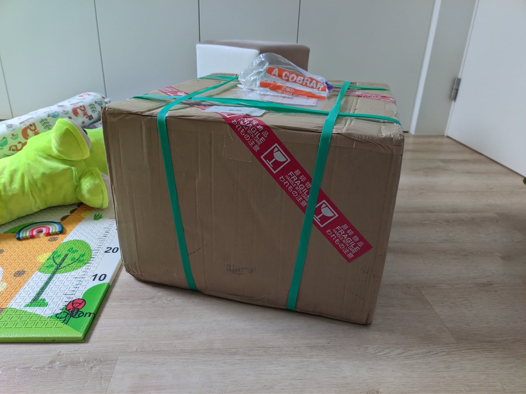 Sealed cardboard shipping box on a wood floor, bound with two green straps and a red diagonal Japanese FRAGILE label, a document pouch on top (the Sony BVM-D9H5J arriving from Japan).