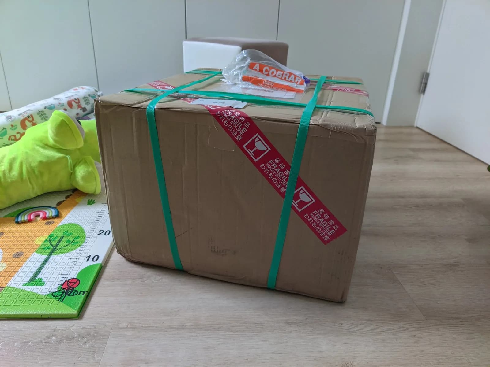 Sealed cardboard shipping box on a wood floor, bound with green straps and a red diagonal Japanese FRAGILE label, a document pouch on top (the Sony BVM-D9H5J and Ikegami CRTs arriving from Japan).