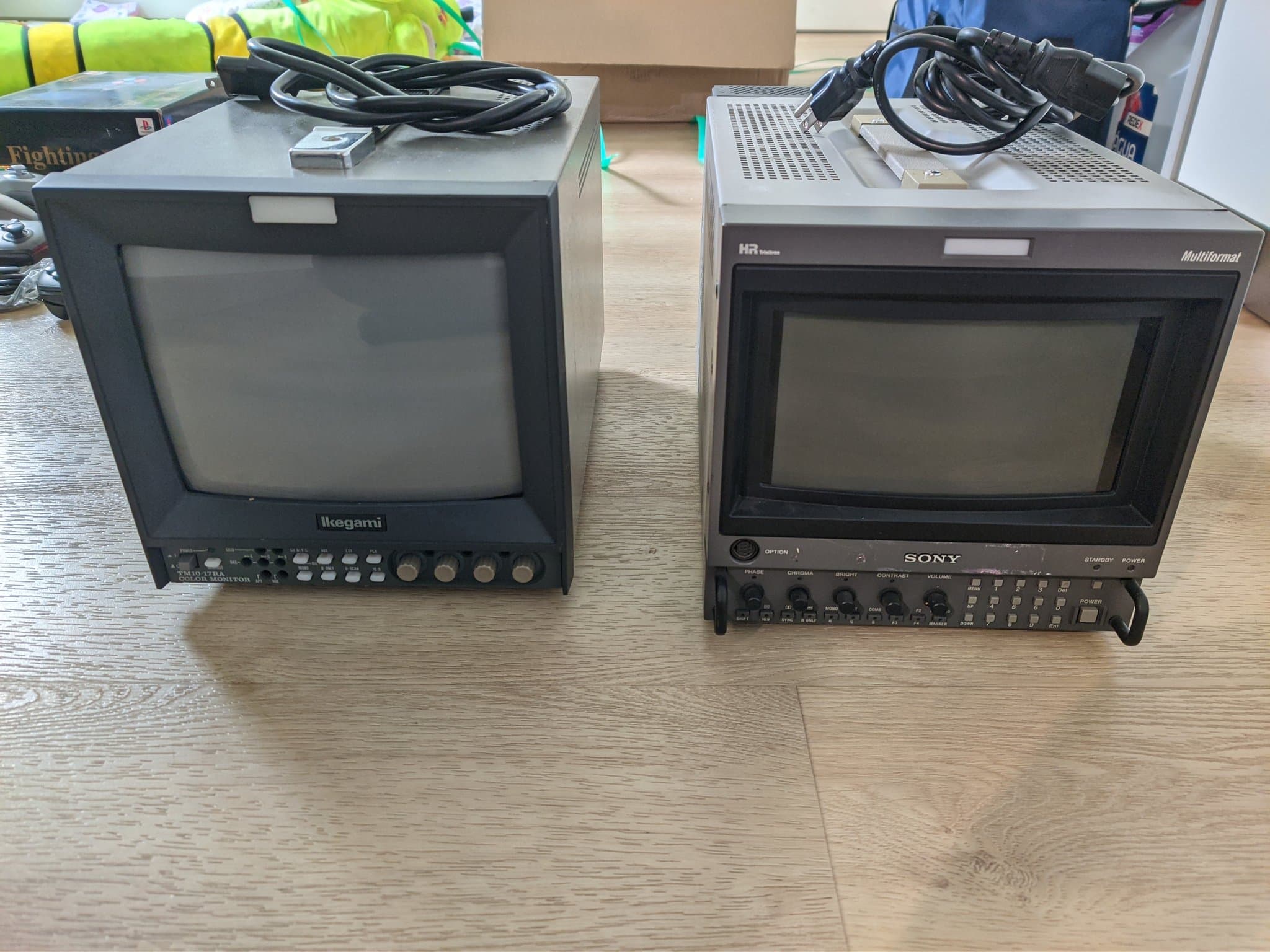 Two broadcast CRTs on a wood desk with blank grey screens: the Ikegami TM10-17RA (left) and the Sony BVM-D9H5J (right, HR Trinitron Multiformat), power cables coiled on top.