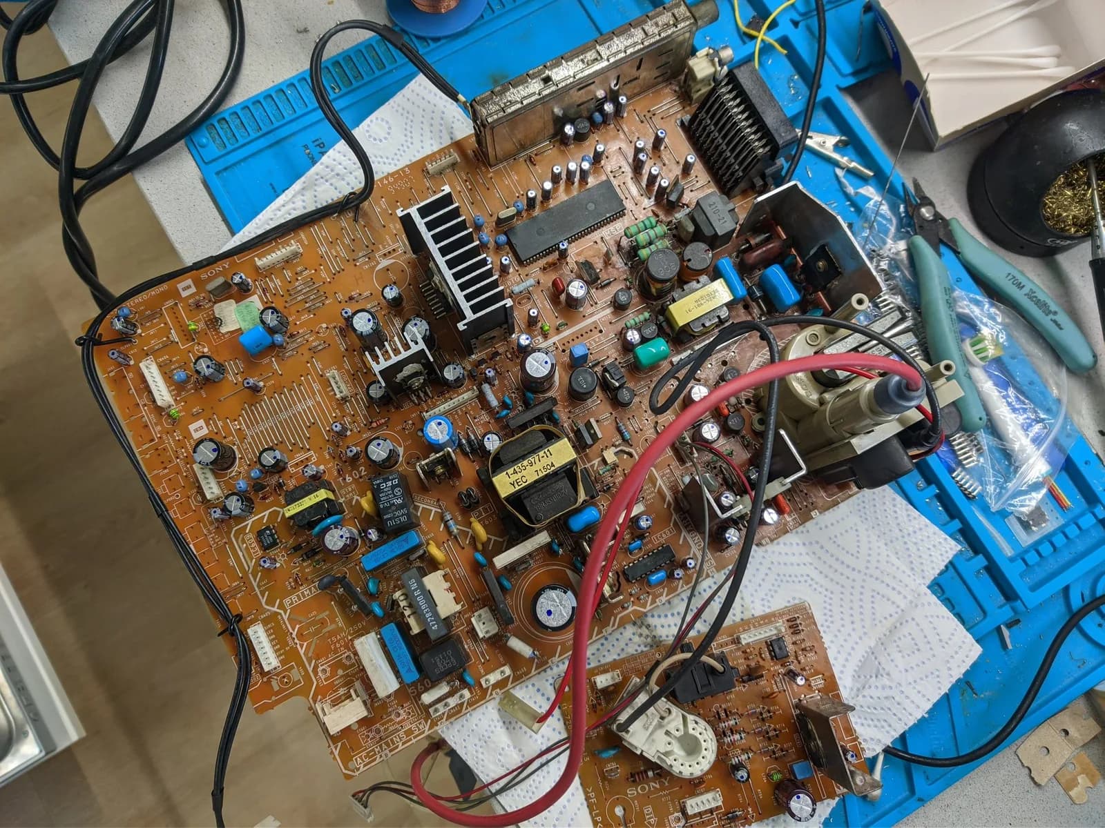 Sony KV-29FX30E main PCB and neck board on a blue silicone mat, both fully recapped with every new capacitor marked by a dot, the heatsink and transformer in place and a soldering iron at the edge.